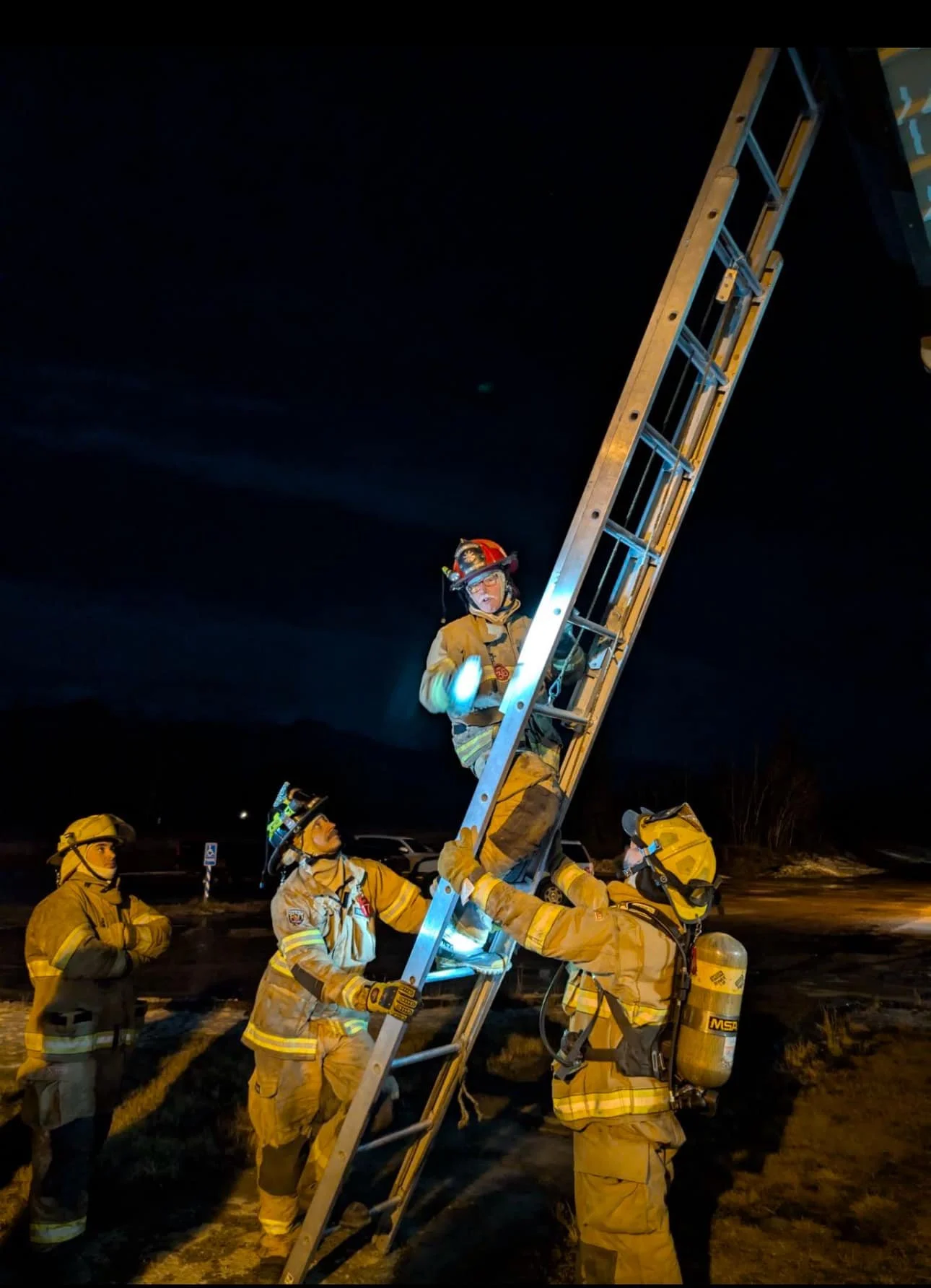 Firefighters in protective gear set up an extension ladder at night, with one firefighter climbing while others support. Dark sky and parking lot in background.