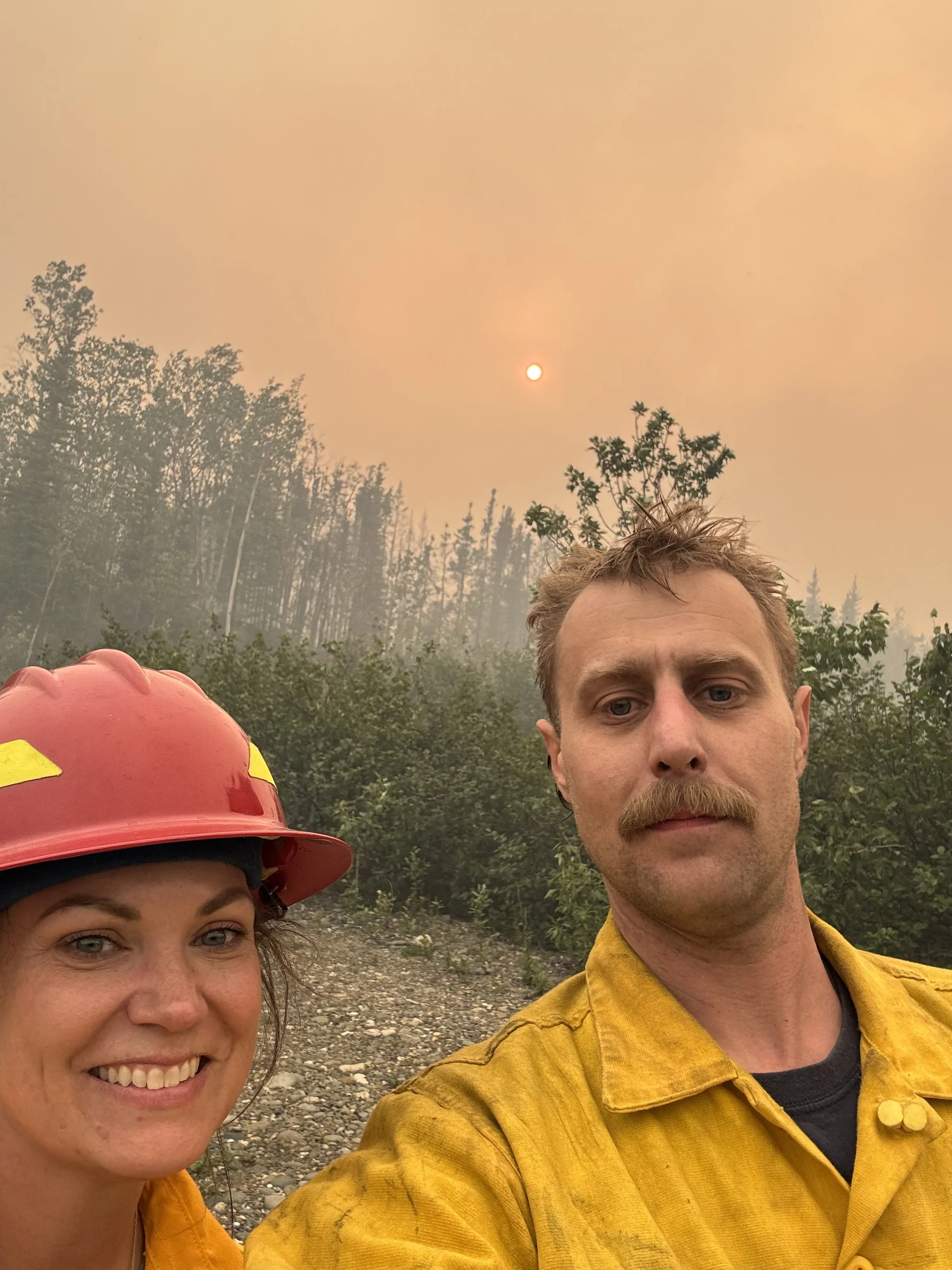 A man and woman taking a selfie outdoors on a smoky day. The man is wearing a yellow fire-resistant jacket and has a mustache and light brown hair. The woman, partially visible, is wearing a red firefighter helmet. In the background, there are trees and a smudged orange sky with the sun visible through the smoke.