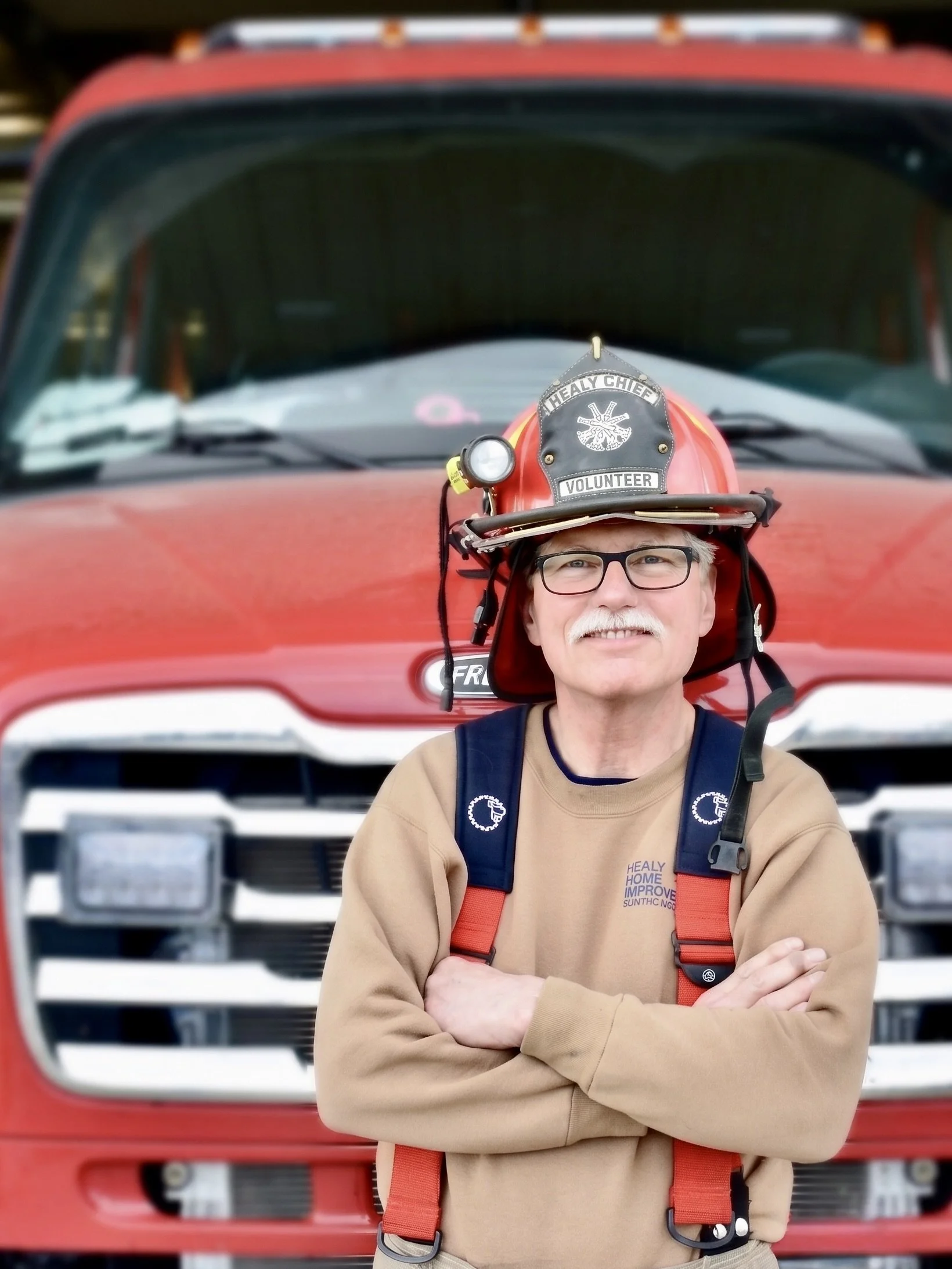 Cartoon boy dressed as a firefighter, wearing a red firefighter suit with yellow accents, a firefighter helmet, and black boots, standing with a confident smile against a light blue background.