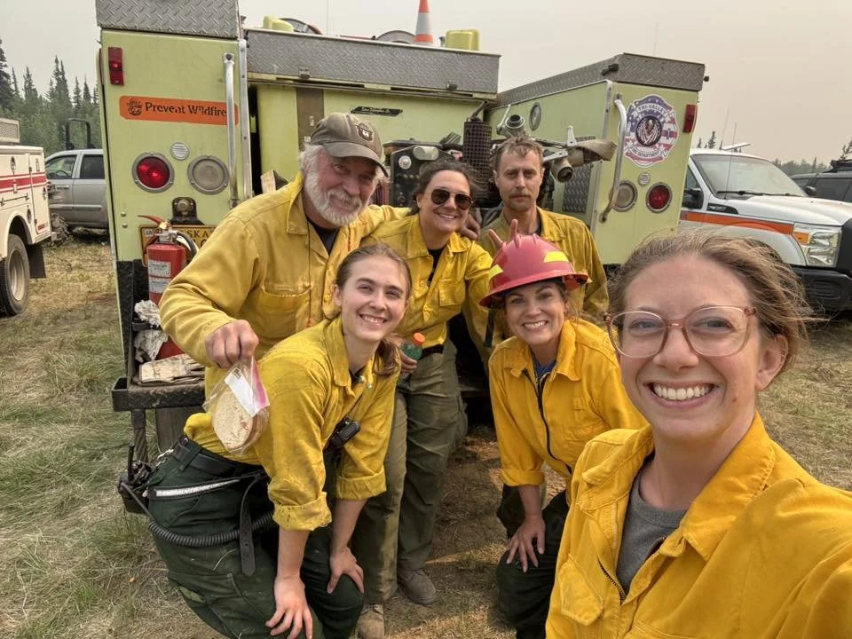 Six firefighters wearing yellow uniforms smiling for a selfie in front of a fire truck with equipment.