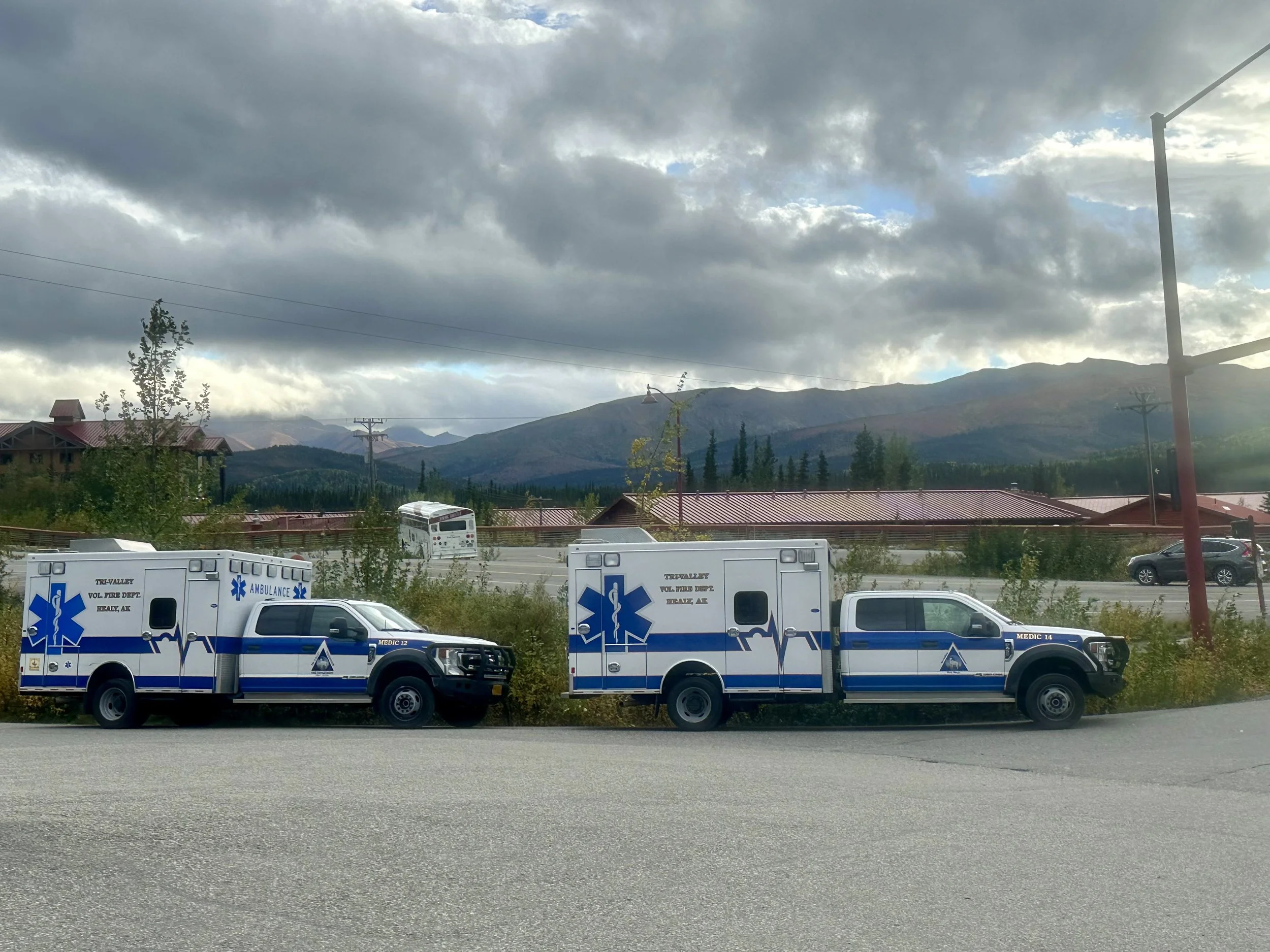 Two ambulances parked in a row on a street, with mountains in the background under a cloudy sky.