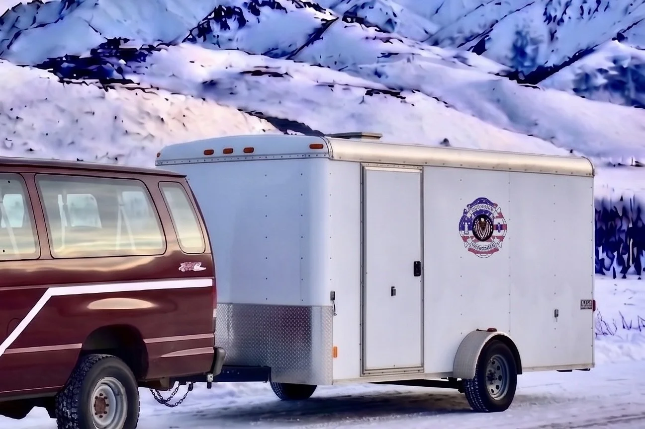 A maroon and white van towing a white enclosed trailer with a logo, parked on a snow-covered road with snow-covered mountains in the background.