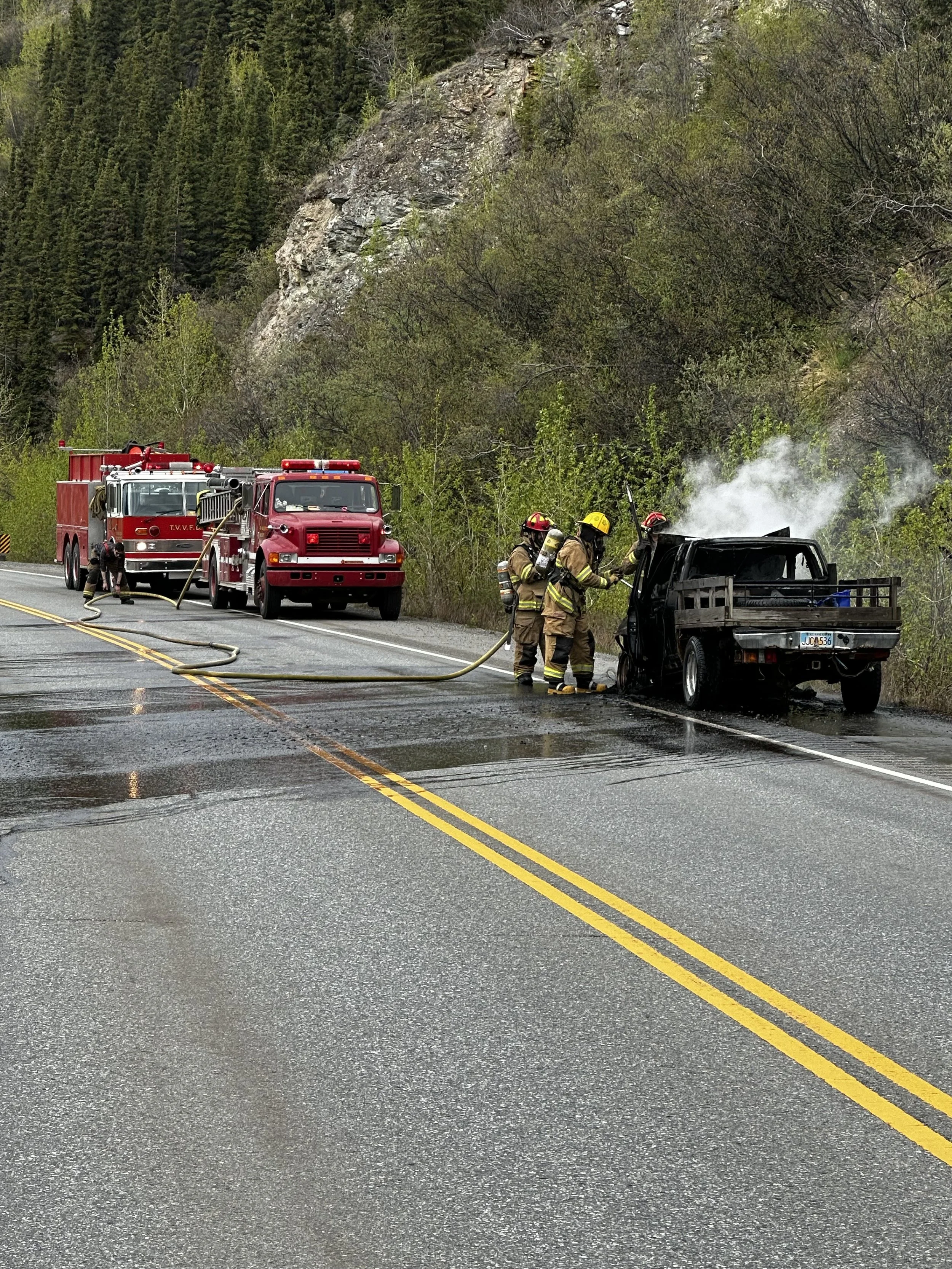 Firefighters extinguishing a car fire on the side of a wet, paved road with a forested hillside in the background.