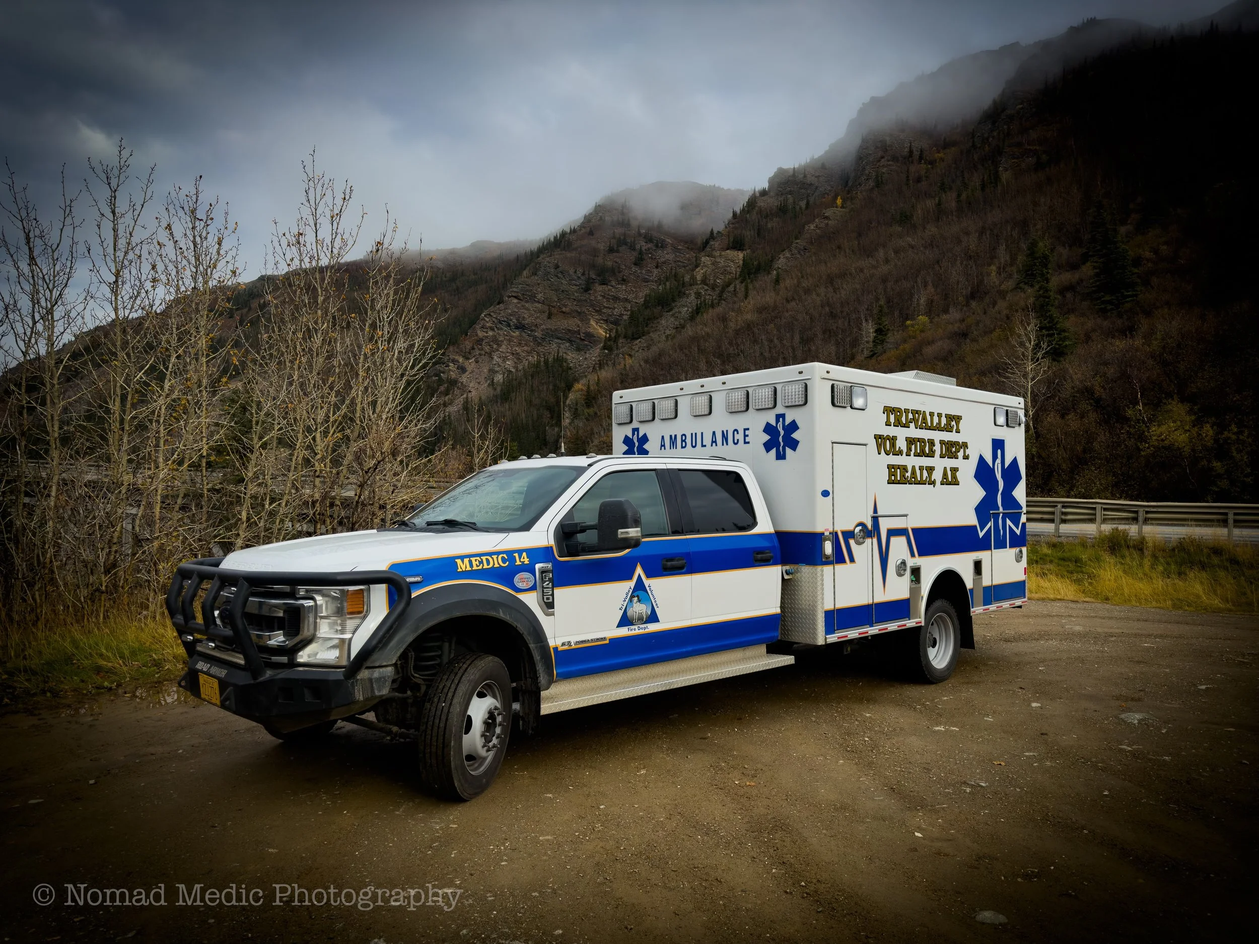 A white ambulance with blue and yellow markings parked on a dirt path with mountainous terrain and fog in the background.