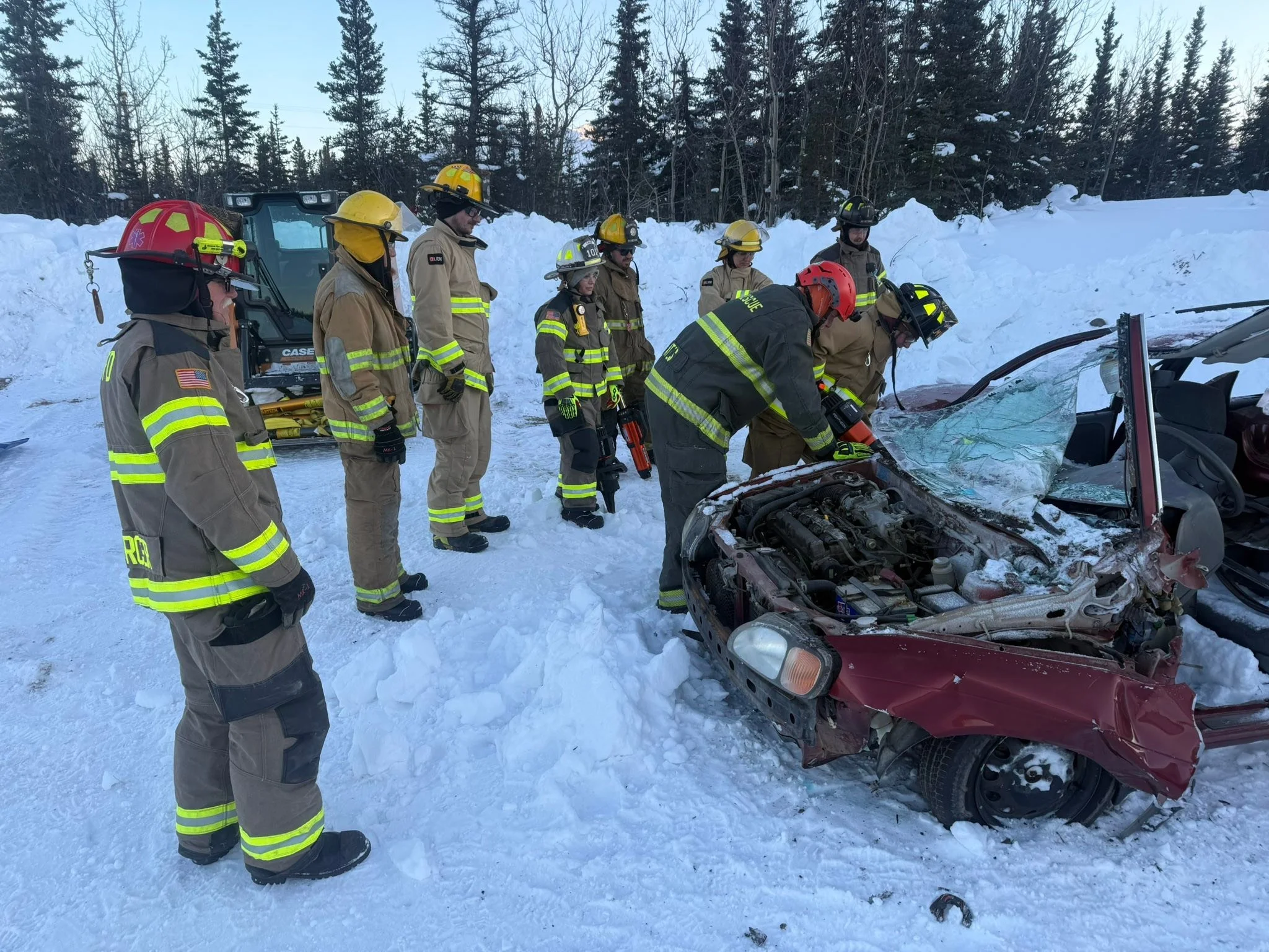 Firefighters and rescue personnel inspecting a car crash site in snowy terrain with severe front-end damage to the vehicle.
