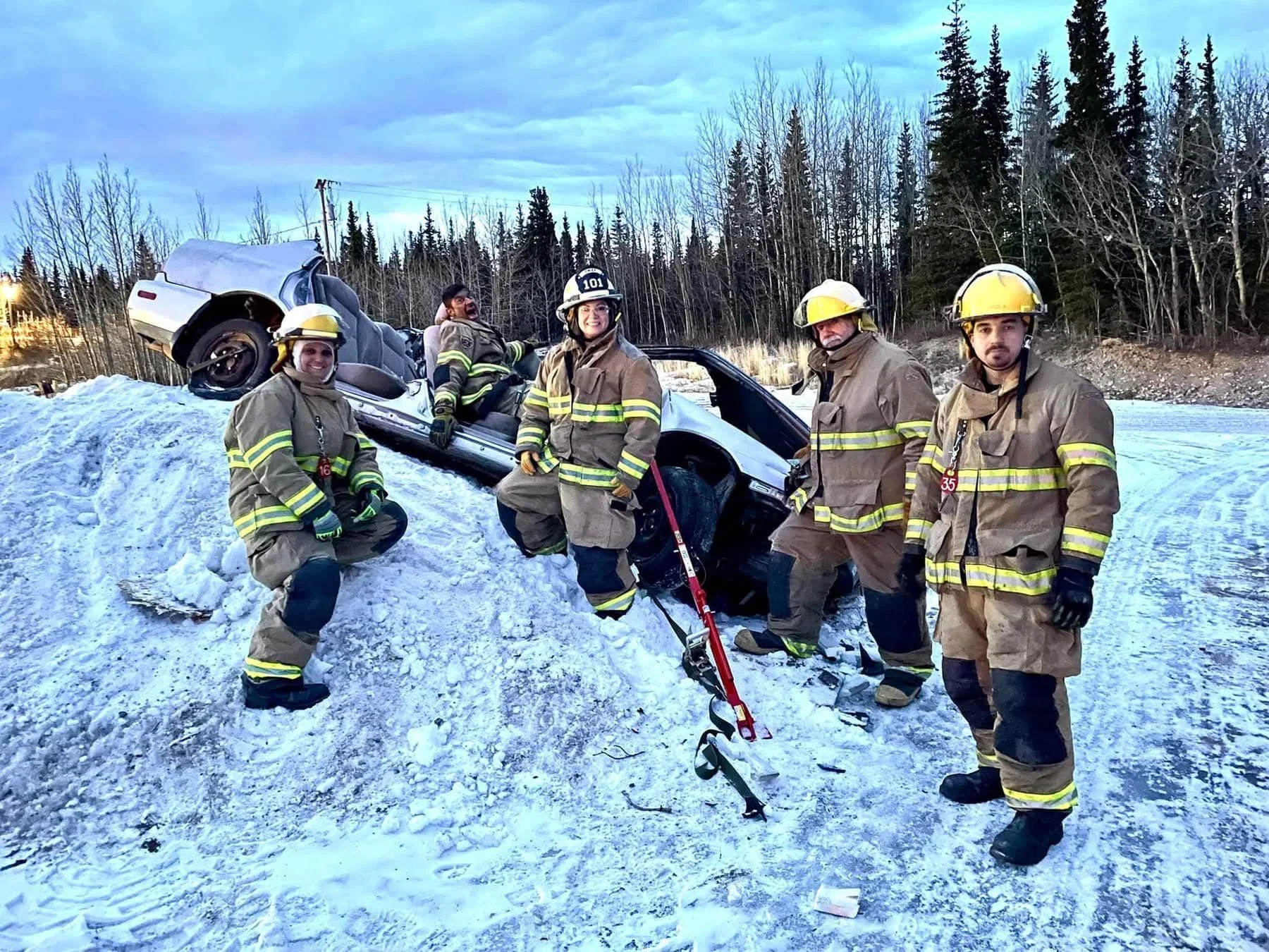 Firefighters working at the scene of a car accident in snowy terrain with a car flipped upside down on a small snow mound and a forest background.