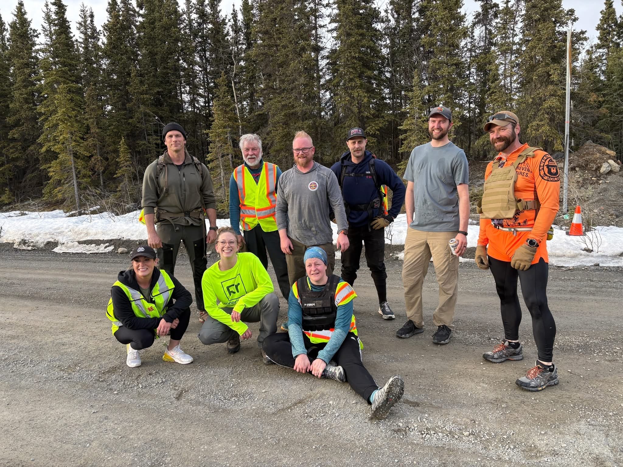 A group of eight people, six men and two women, standing on a snowy road in a forested area. They are dressed in outdoor or safety gear, including vests, jackets, and hats, and are smiling or looking relaxed.