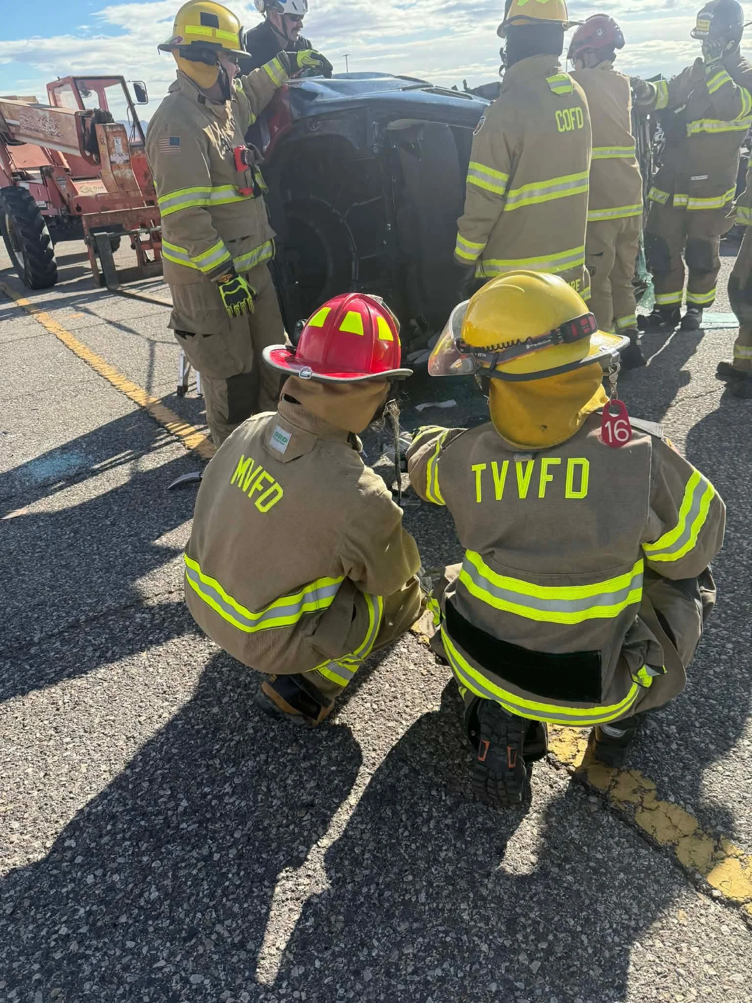 Firefighters from TUVFD, MIFD, and COFD wearing protective gear and helmets, gathered around a damaged black vehicle on a road, with rescue equipment nearby, under a cloudy sky.