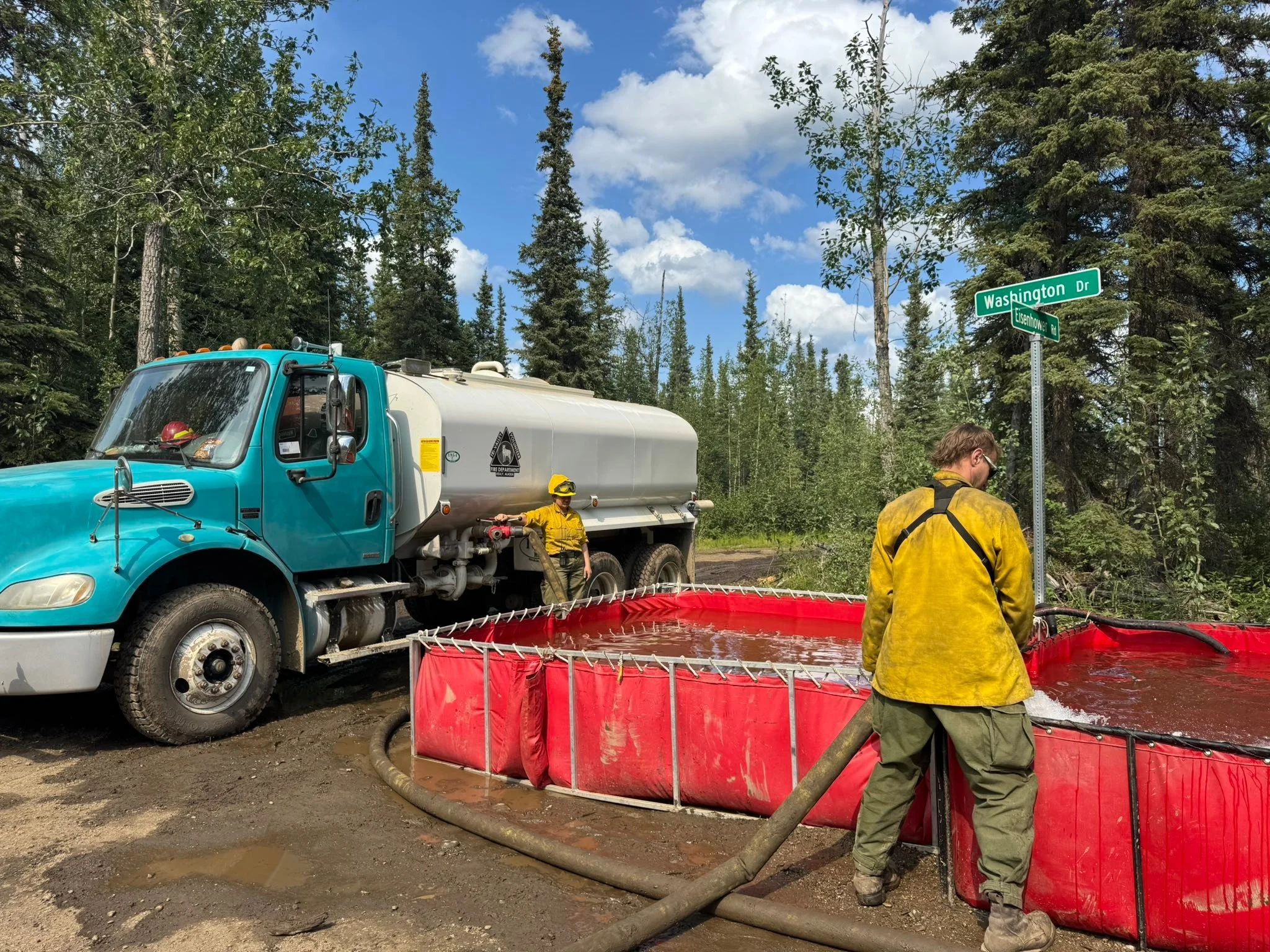 Firefighters or workers in yellow protective gear work near a large blue water truck and a red containment pool filled with muddy water, on a dirt road surrounded by tall trees under a partly cloudy sky.