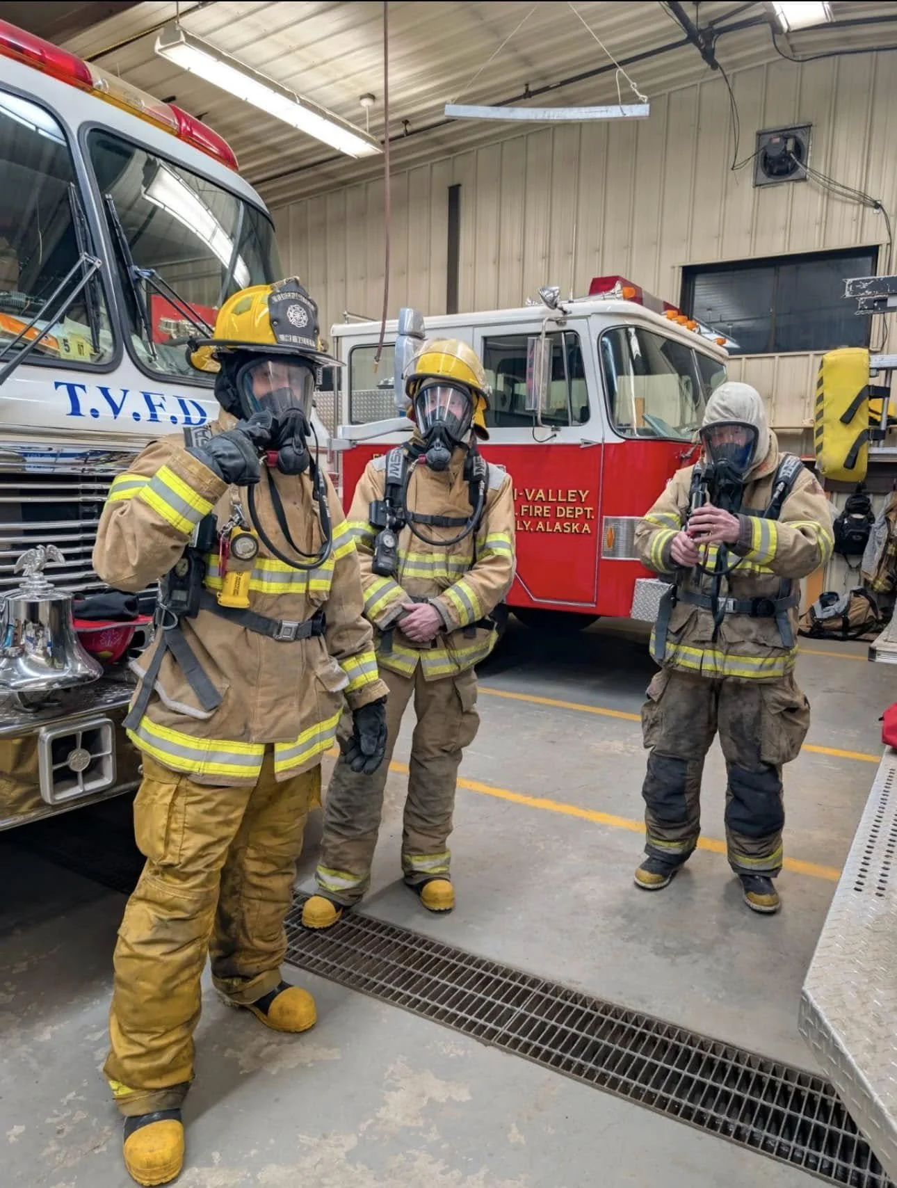 Three firefighters in protective gear and masks inside a fire station, standing next to fire trucks.