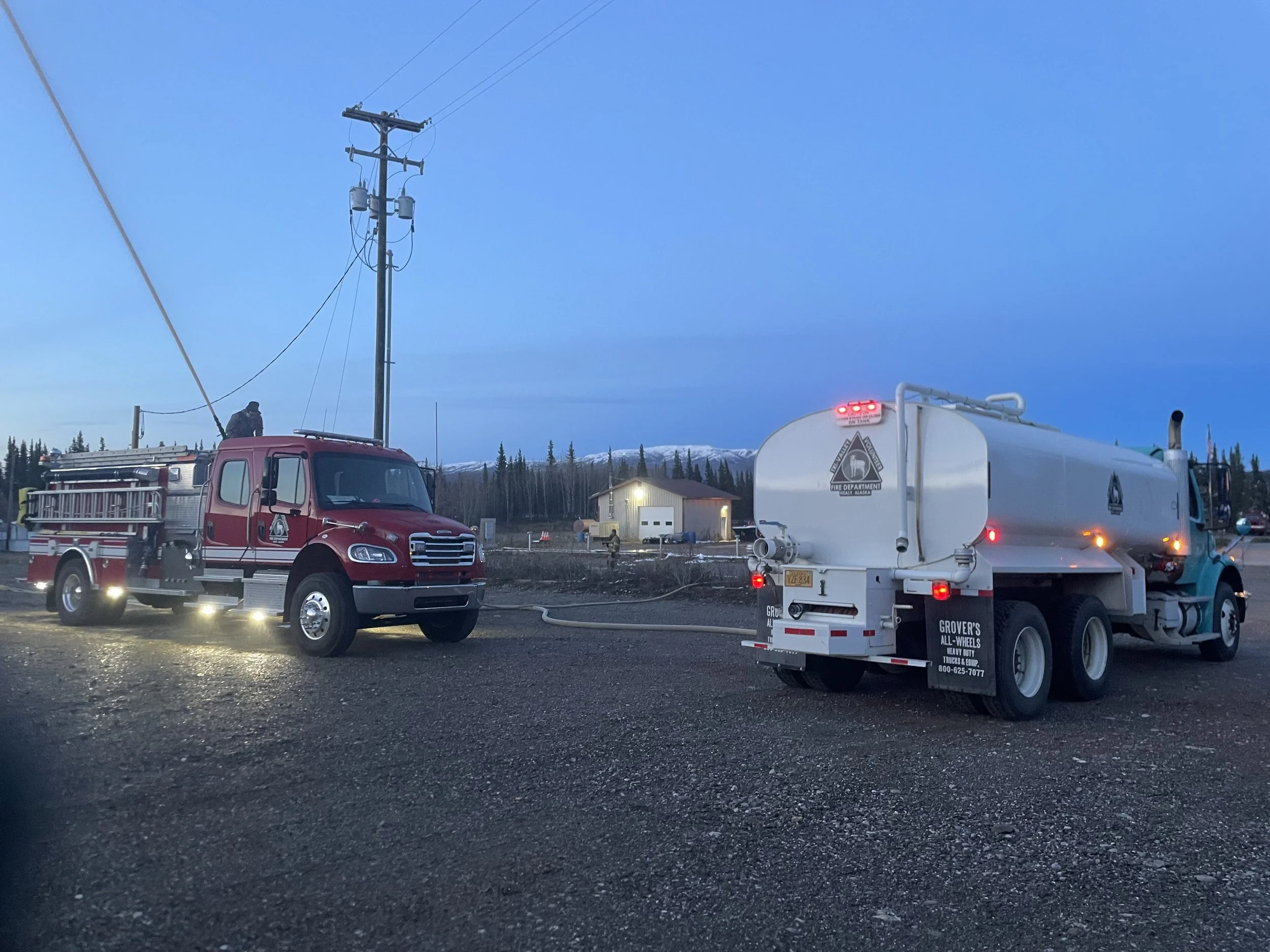A scene with emergency vehicles, including a red fire truck and a white tanker truck, parked on a dark gravel lot near a building. There are power lines overhead, and snow-capped mountains and trees are visible in the background during dusk or dawn.