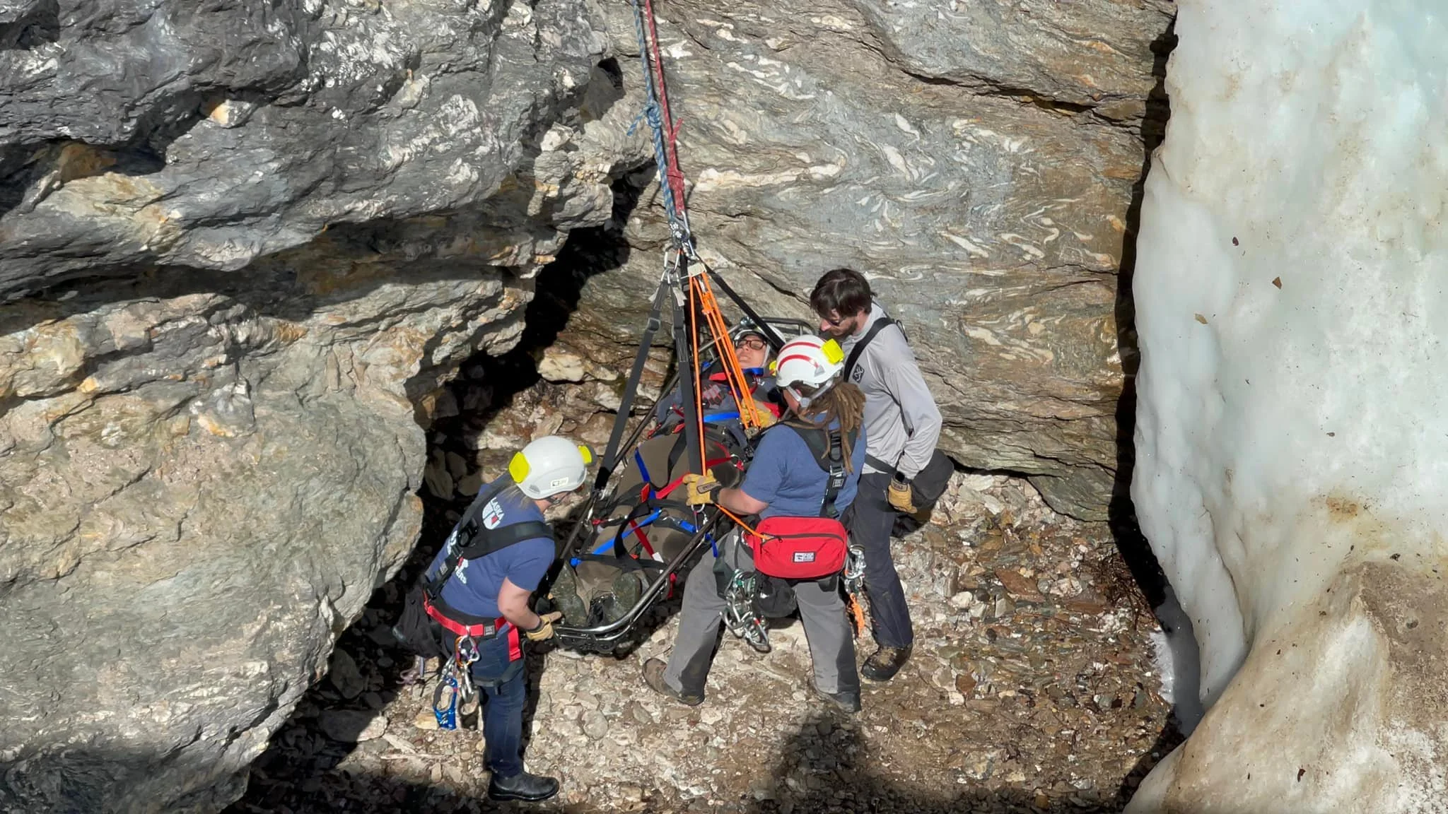 Rescue team members in outdoor gear lifting a person on a stretcher through a narrow, rocky crevice between mountain cliffs.