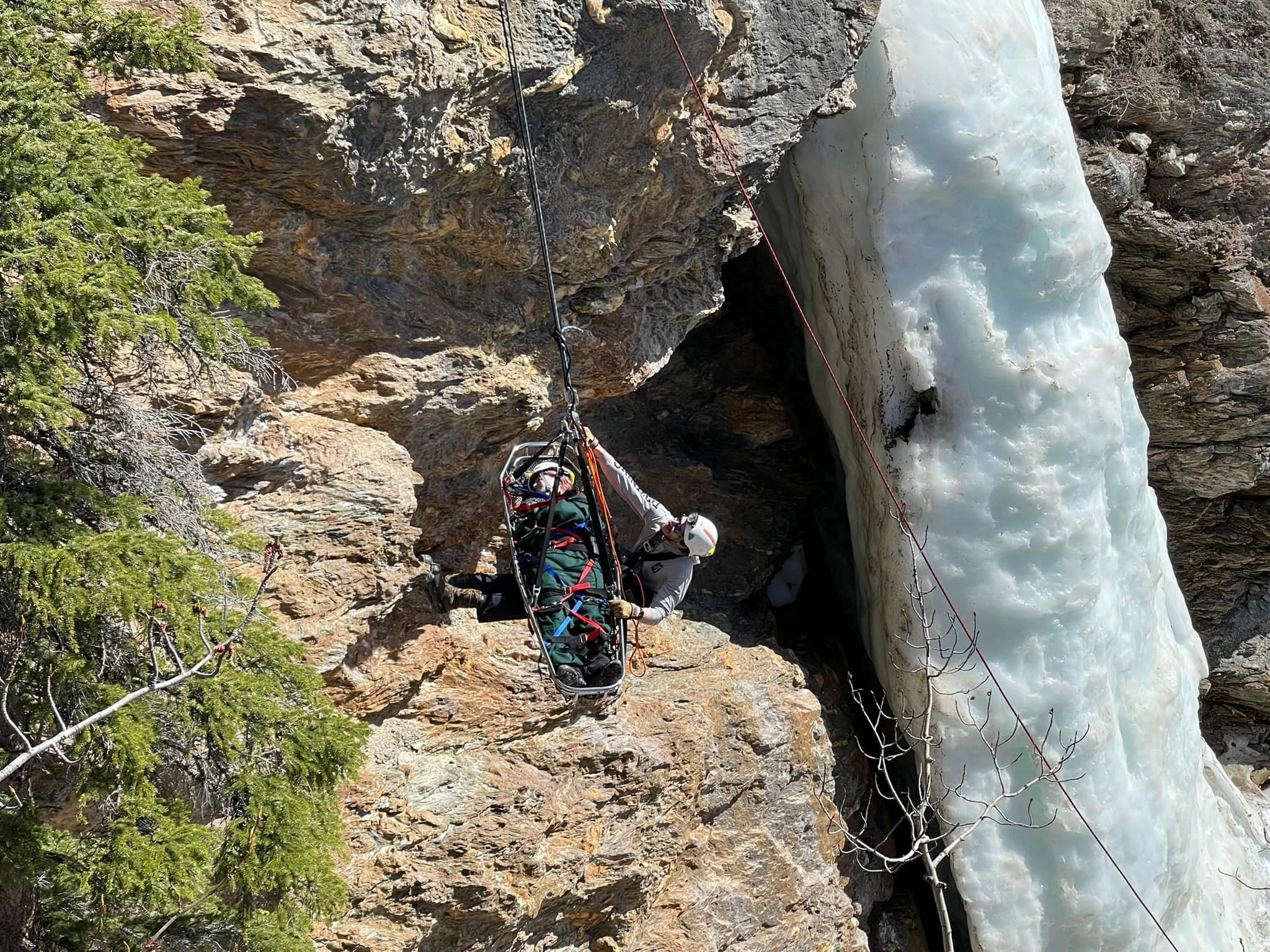 A person in a white helmet and outdoor gear is lying in a rescue stretcher near a rocky cliffside with ice and water, possibly ice climbing or rescue operation.