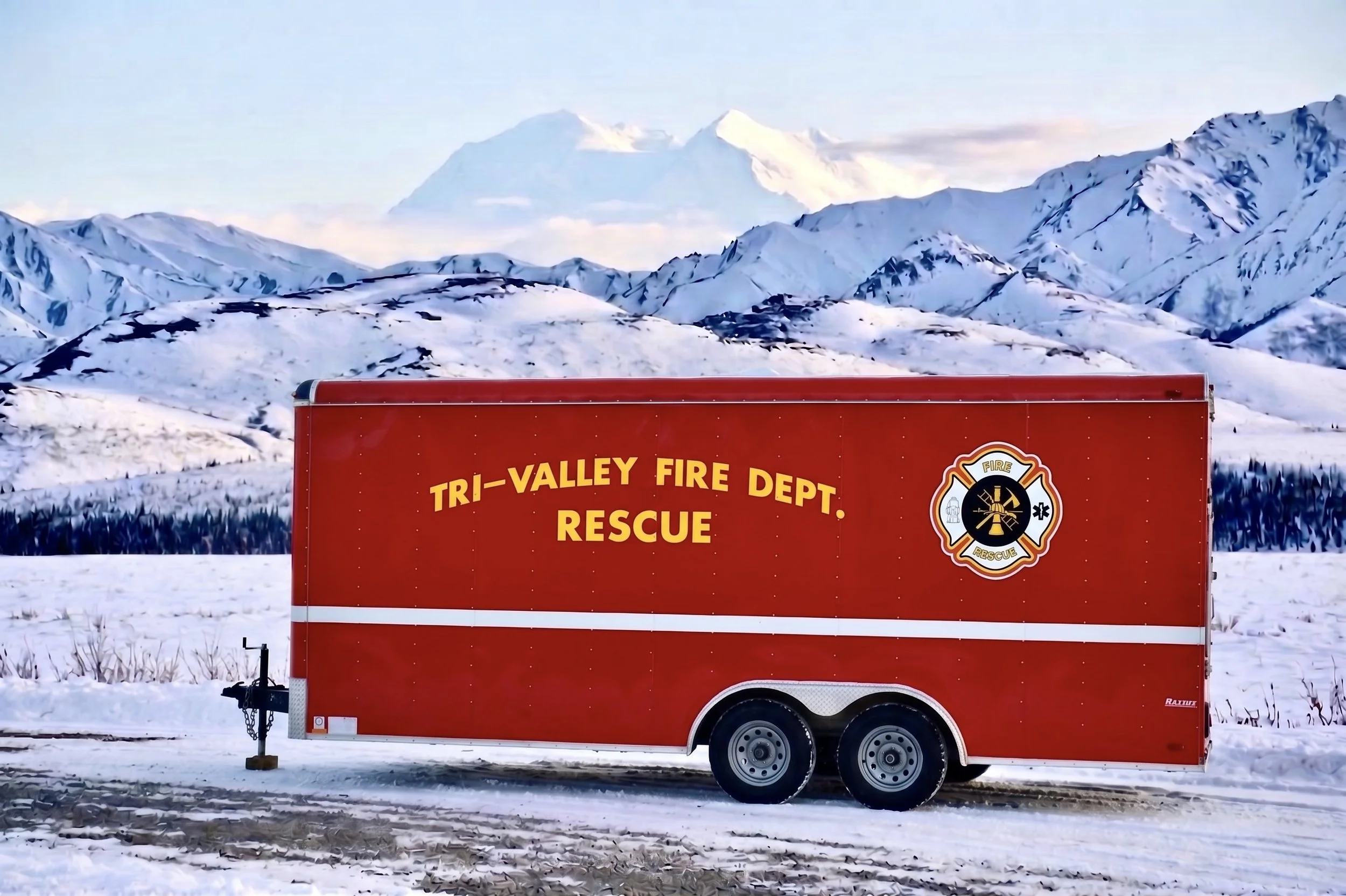 A red rescue trailer from Tri-Valley Fire Department in a snowy landscape with snow-covered mountains and a partly cloudy sky in the background.