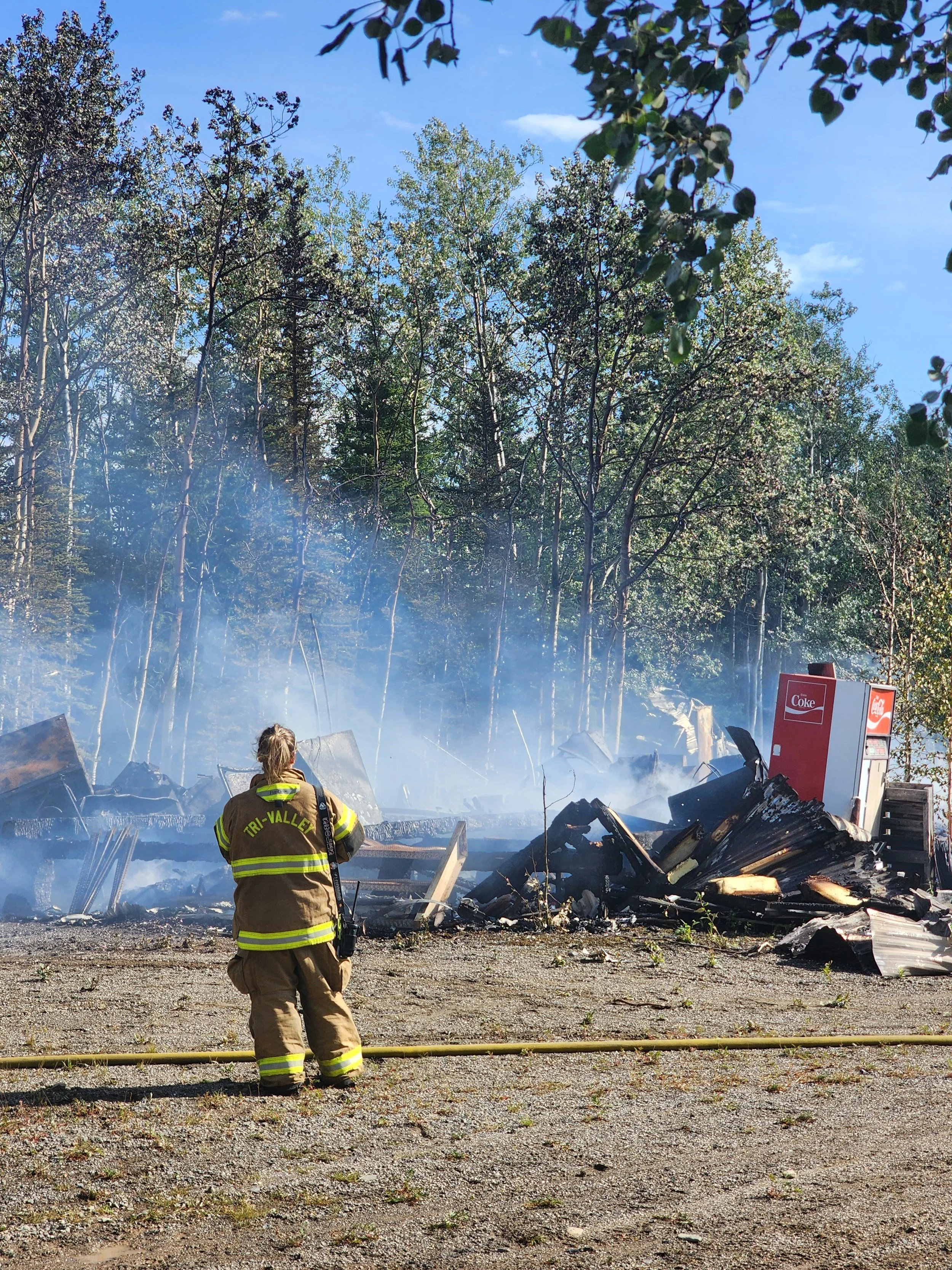 Firefighter in tan uniform with reflective stripes and the words "TRI-VALLEY" on the back, facing burned debris and smoldering remains from a fire, in front of a cloudy sky and trees, with a red Coke vending machine on the right.