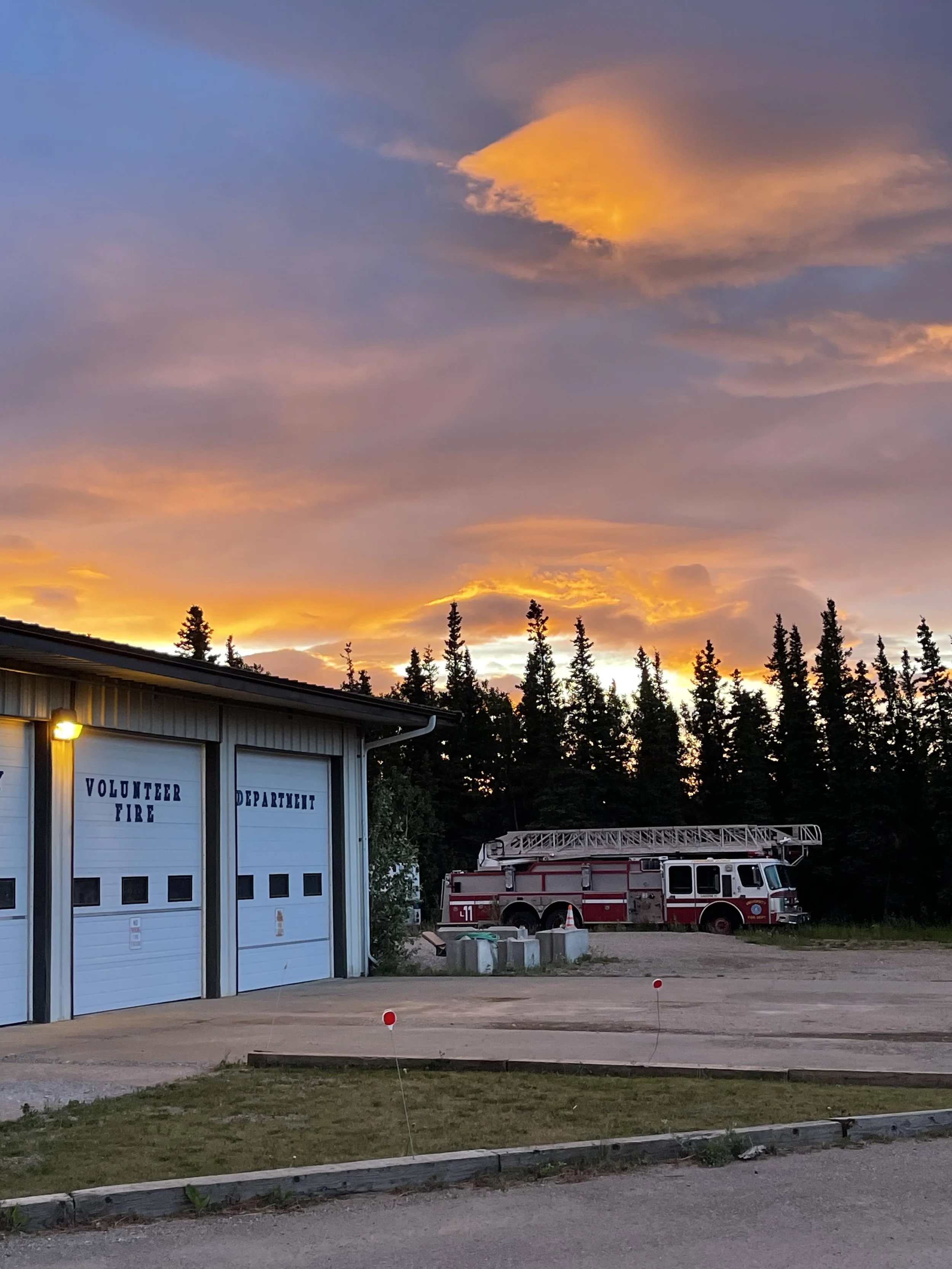 A fire truck parked outside a fire station during sunset with a sky filled with colorful clouds and a row of trees in the background.