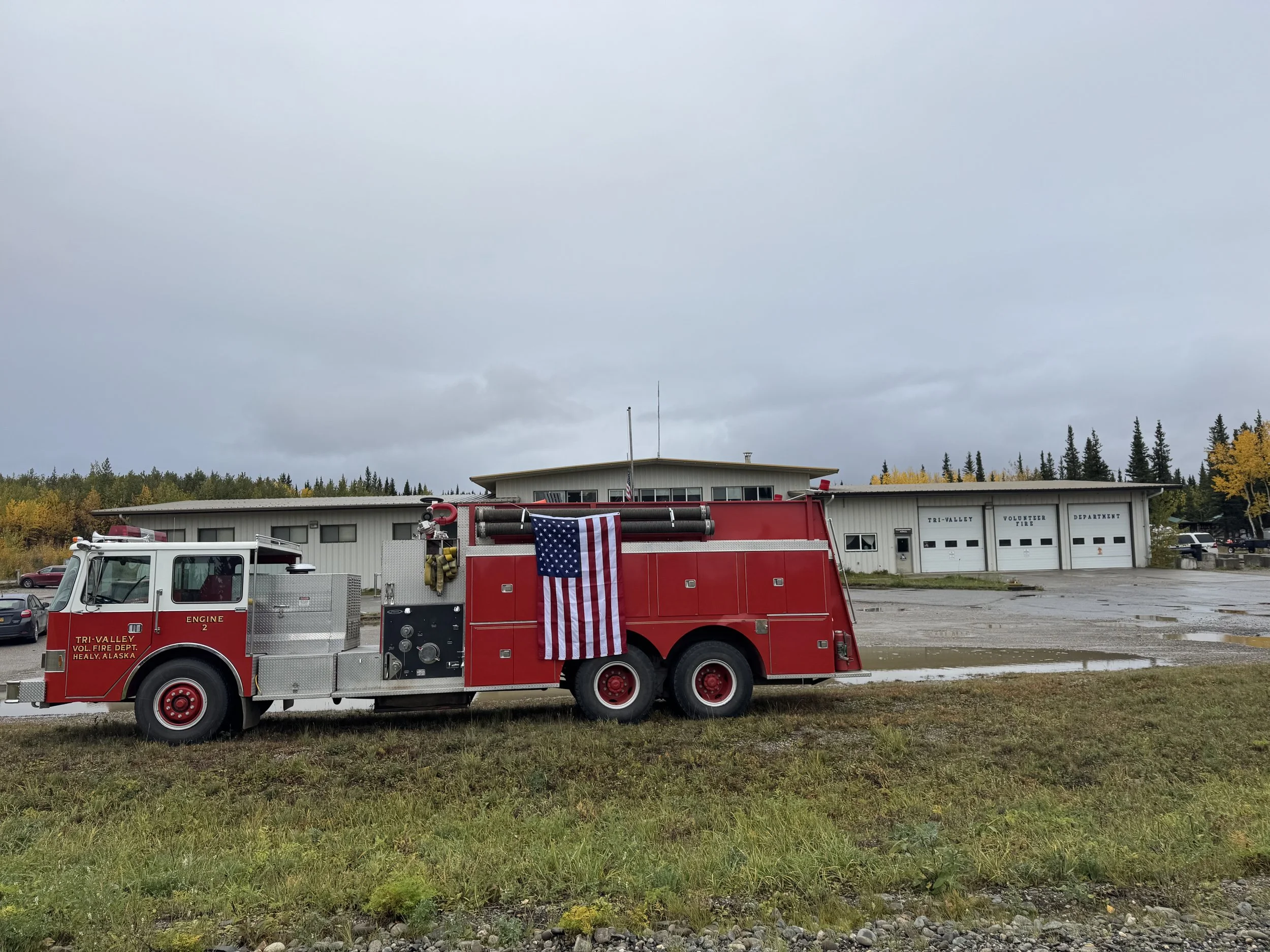 A red fire truck parked on grass in front of a building with a sign reading TRI-VALUE VOLUNTEER FIRE DEPARTMENT, Healy, Alaska. The sky is overcast, and there are trees with fall-colored leaves in the background.