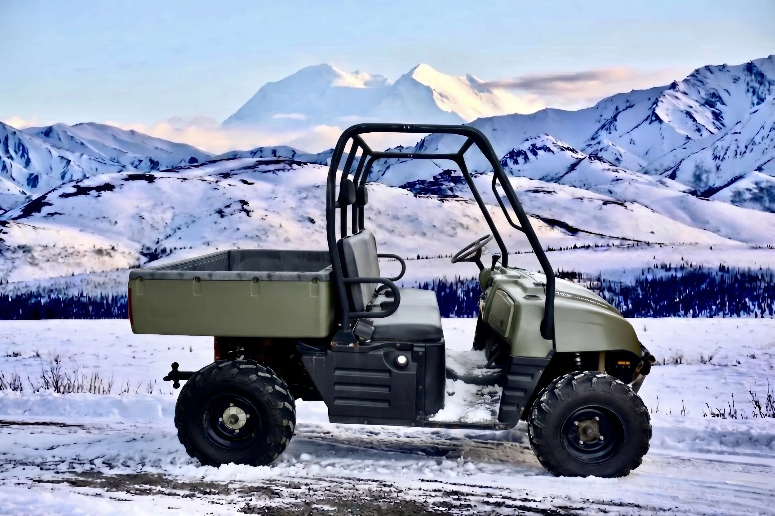 A utility vehicle on snow-covered ground with snow-capped mountains and a blue sky in the background.