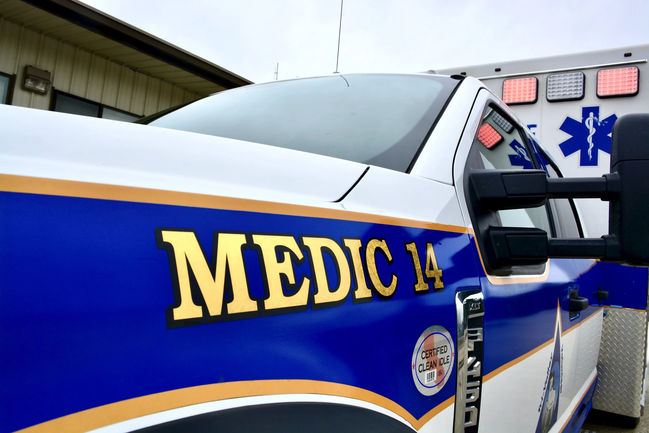Close-up of emergency medical services vehicle with 'MEDIC 14' printed on the side and a blue medical symbol with a staff and snake. An ambulance is partially visible in the background.