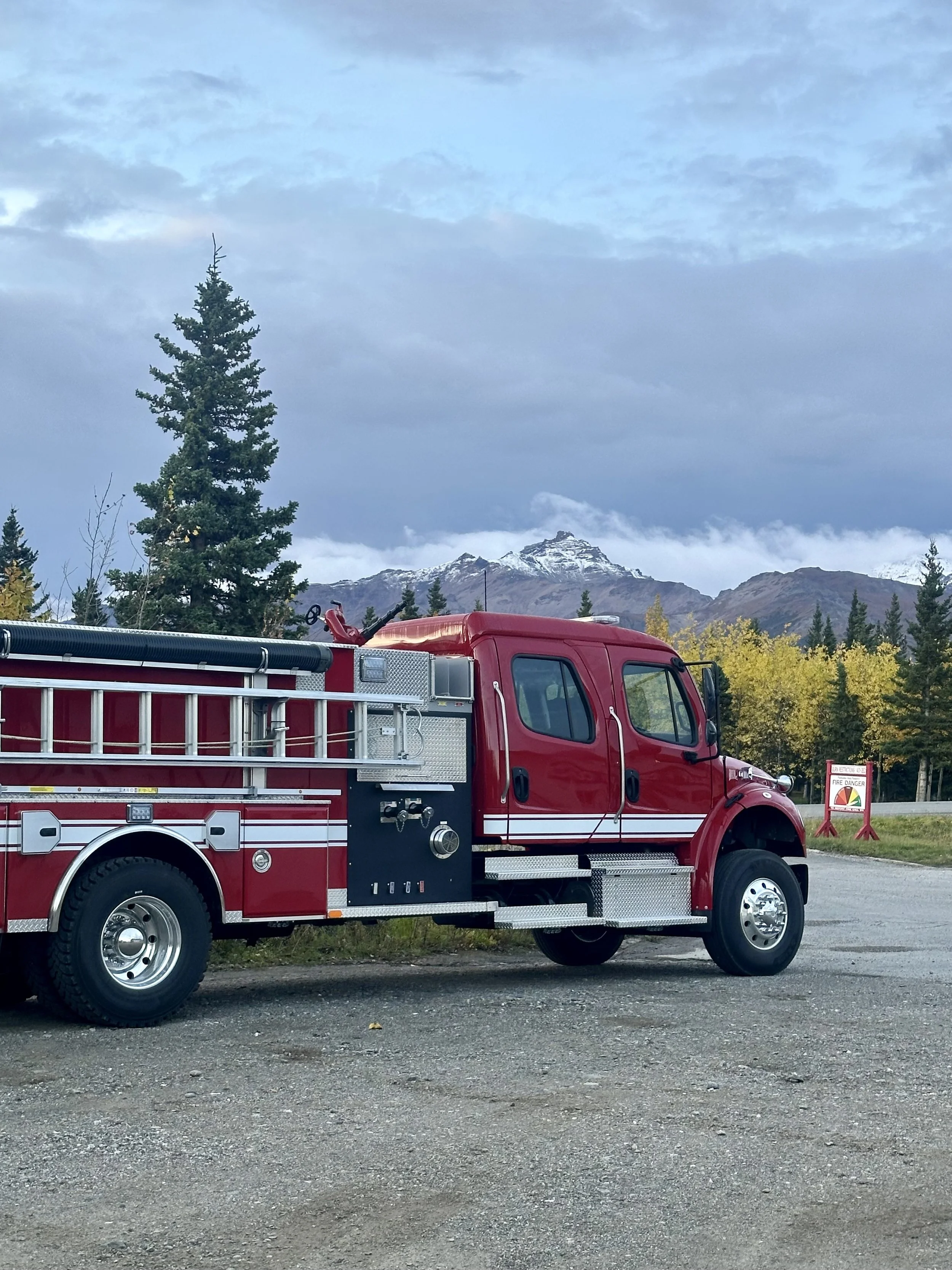 Red fire truck parked on gravel with a background of evergreen trees, mountains with snow, and a partly cloudy sky.