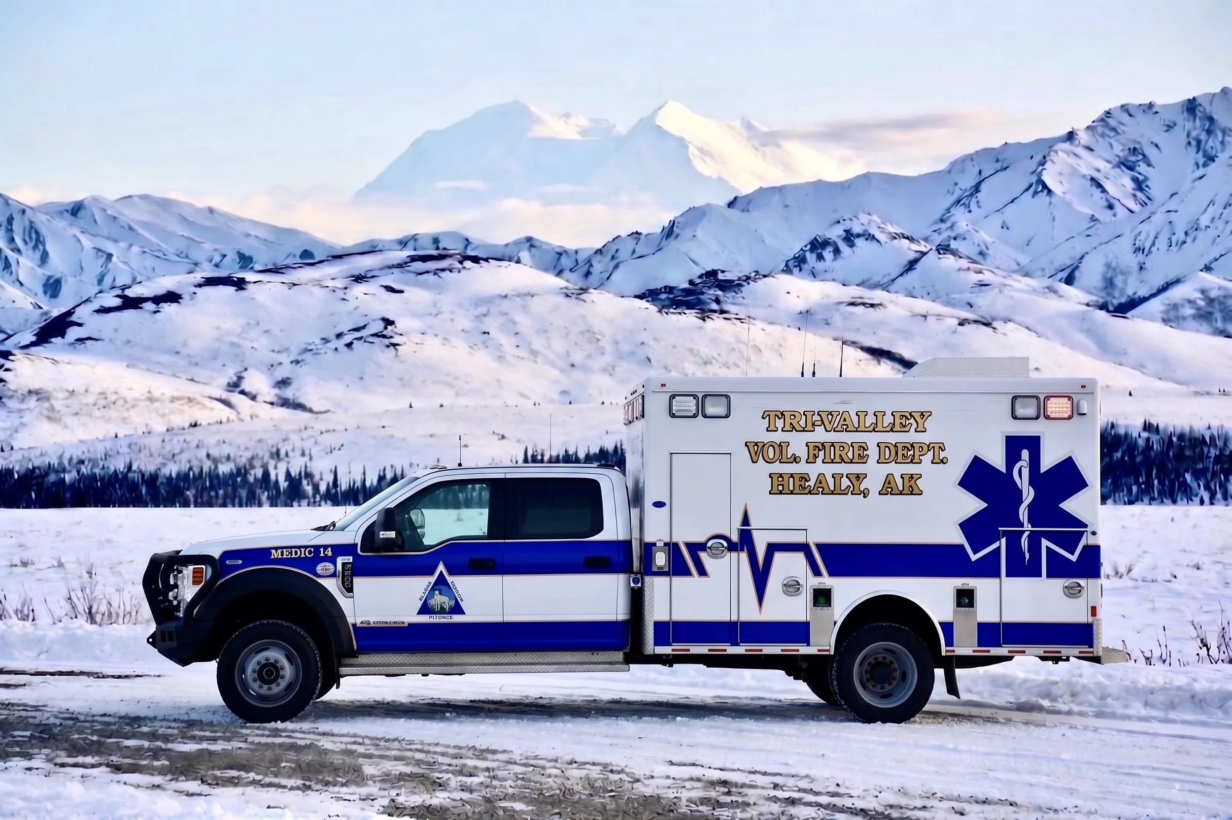 An ambulance parked on snow-covered ground in a mountainous area with snow-capped peaks in the background.
