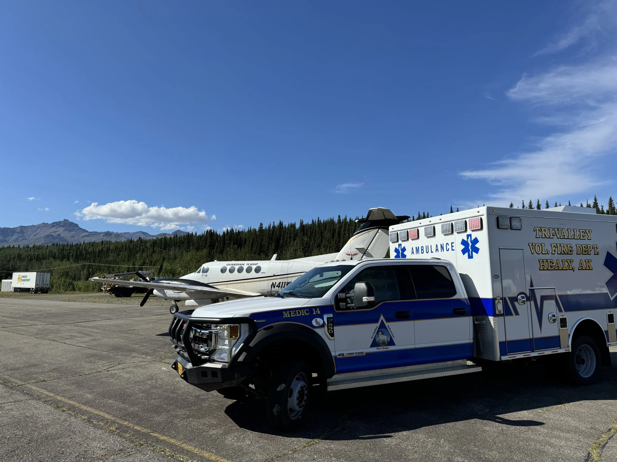 A parking lot with emergency service vehicles including an ambulance and a medical helicopter, with mountains and trees in the background under a blue sky with some clouds.