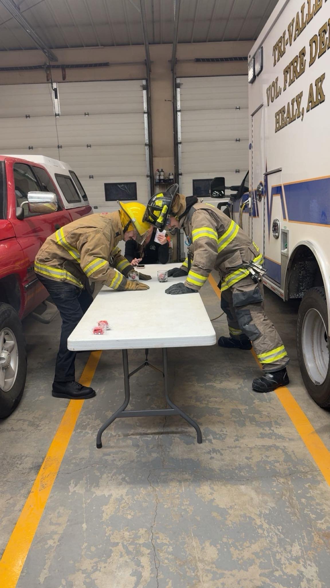 Two firefighters in full gear, including helmets, are leaning over a table inside a fire station. They appear to be examining or discussing something on the table. A person in a dark jacket is sitting at the table and holding a phone. Behind them, there is a red pickup truck and a large fire truck. The fire station has a concrete floor with yellow parking lines and large garage doors.