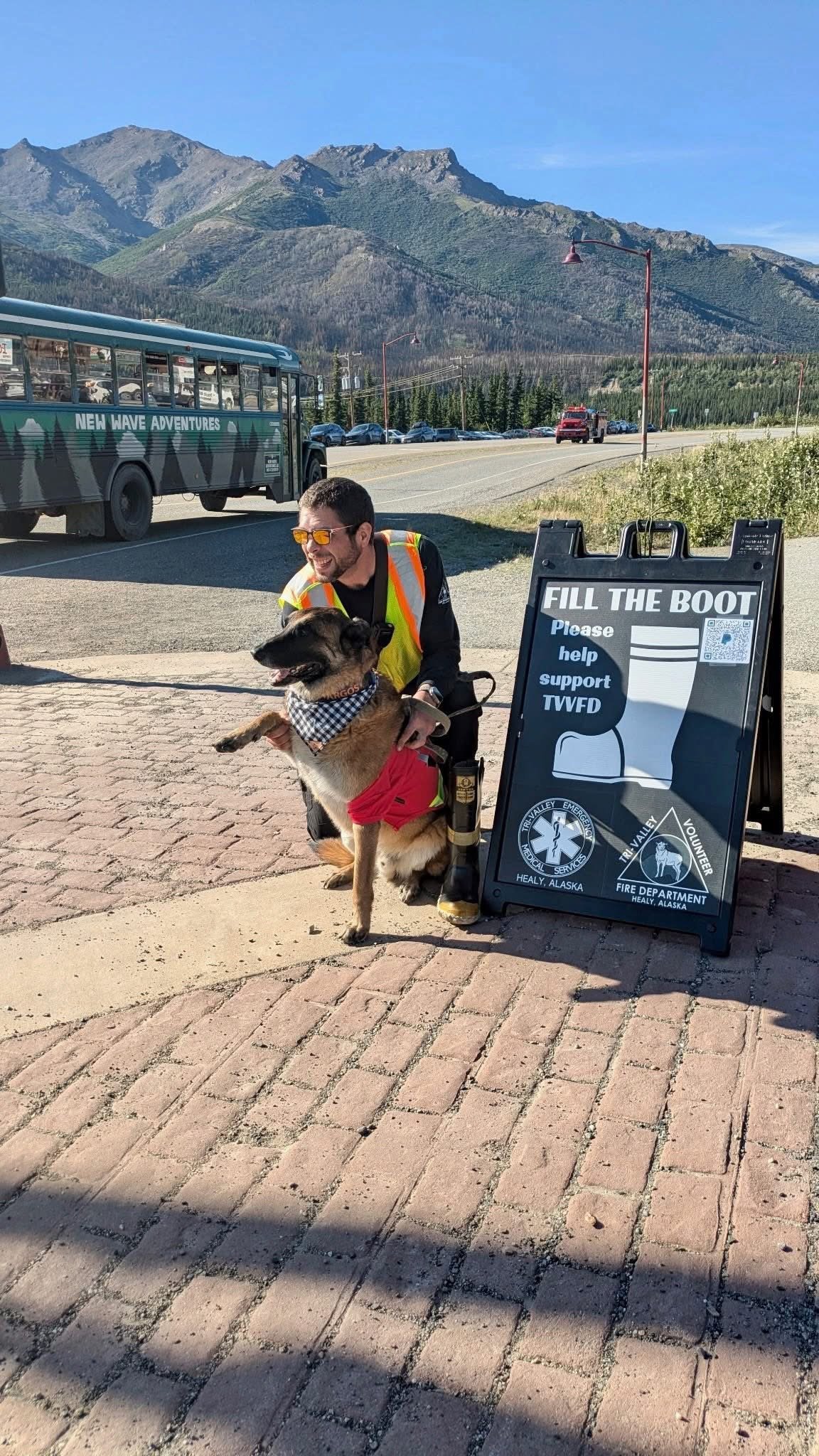 A man wearing a yellow safety vest and sunglasses kneels beside a dog dressed in a red vest. They are next to a sign that encourages filling the boots to support the TVFD and local emergency services in Healy, Alaska. A mountain landscape with a bus, cars, and clear blue sky is in the background.