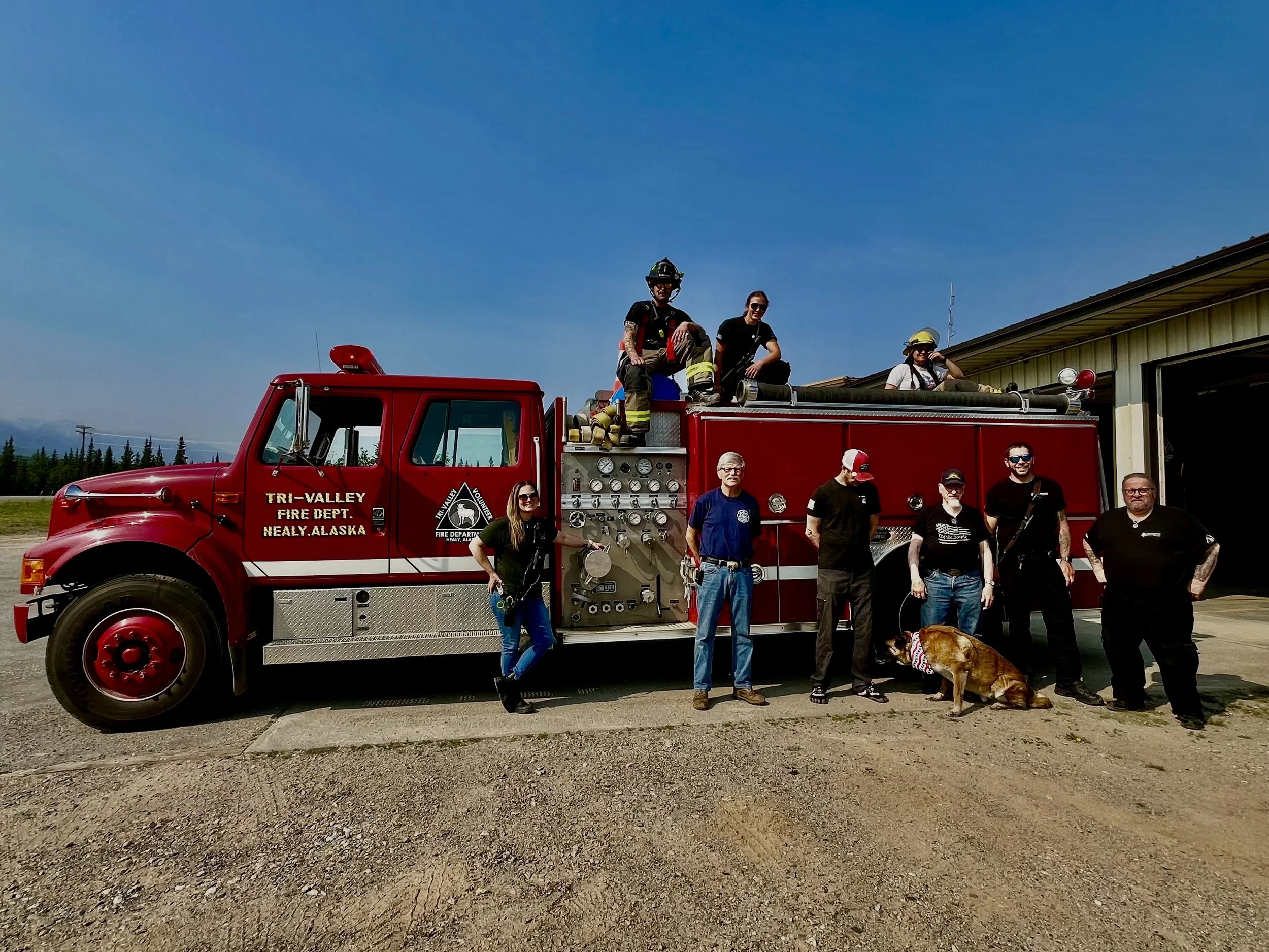 People standing around and sitting on a red fire truck labeled "Tri-Valley Fire Dept. Healy, Alaska," with firefighters and civilians, and a park ranger in uniform, under a clear blue sky.