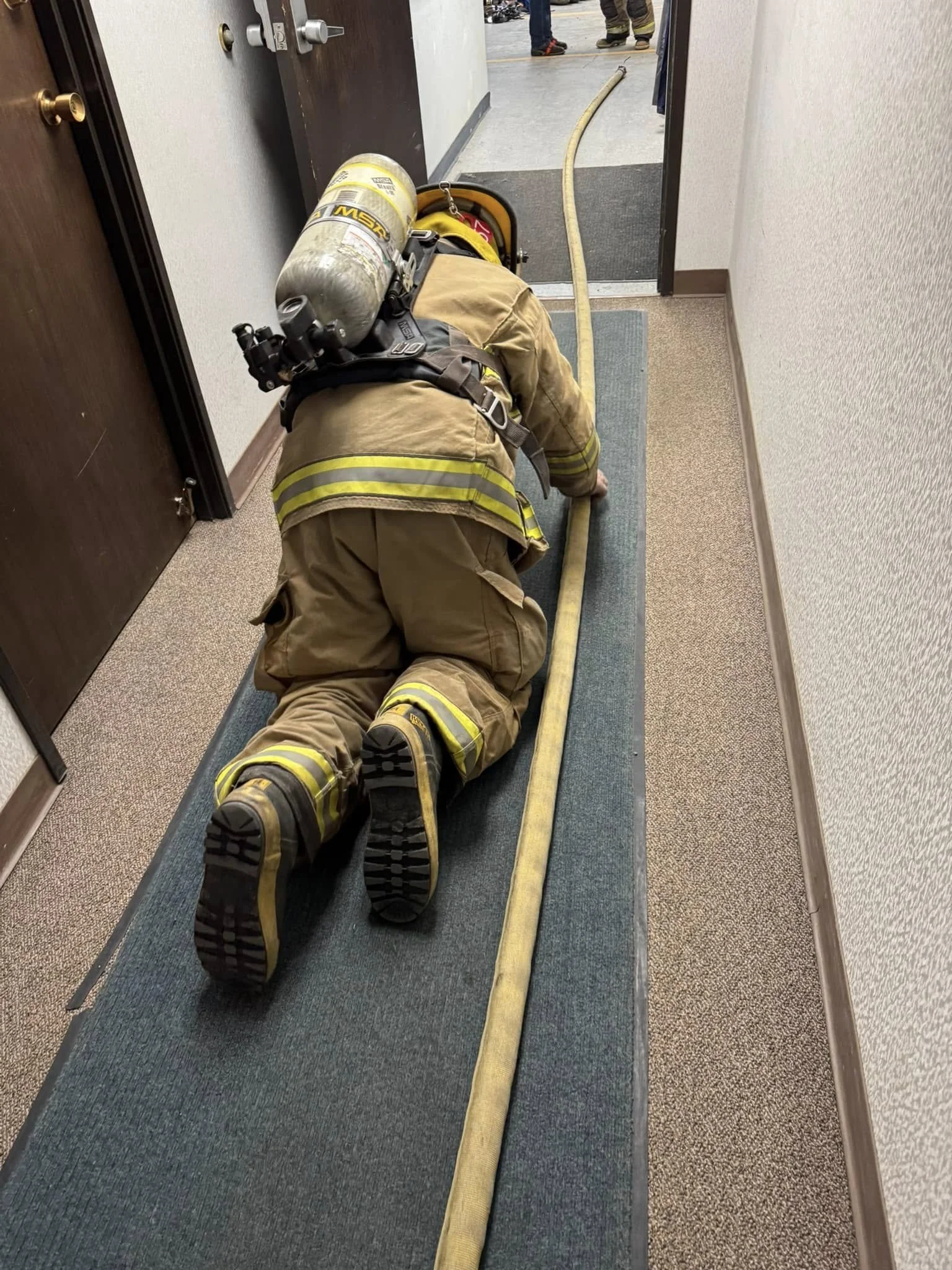 Firefighter crawling on the carpeted floor along a yellow hose in a corridor near an open door.