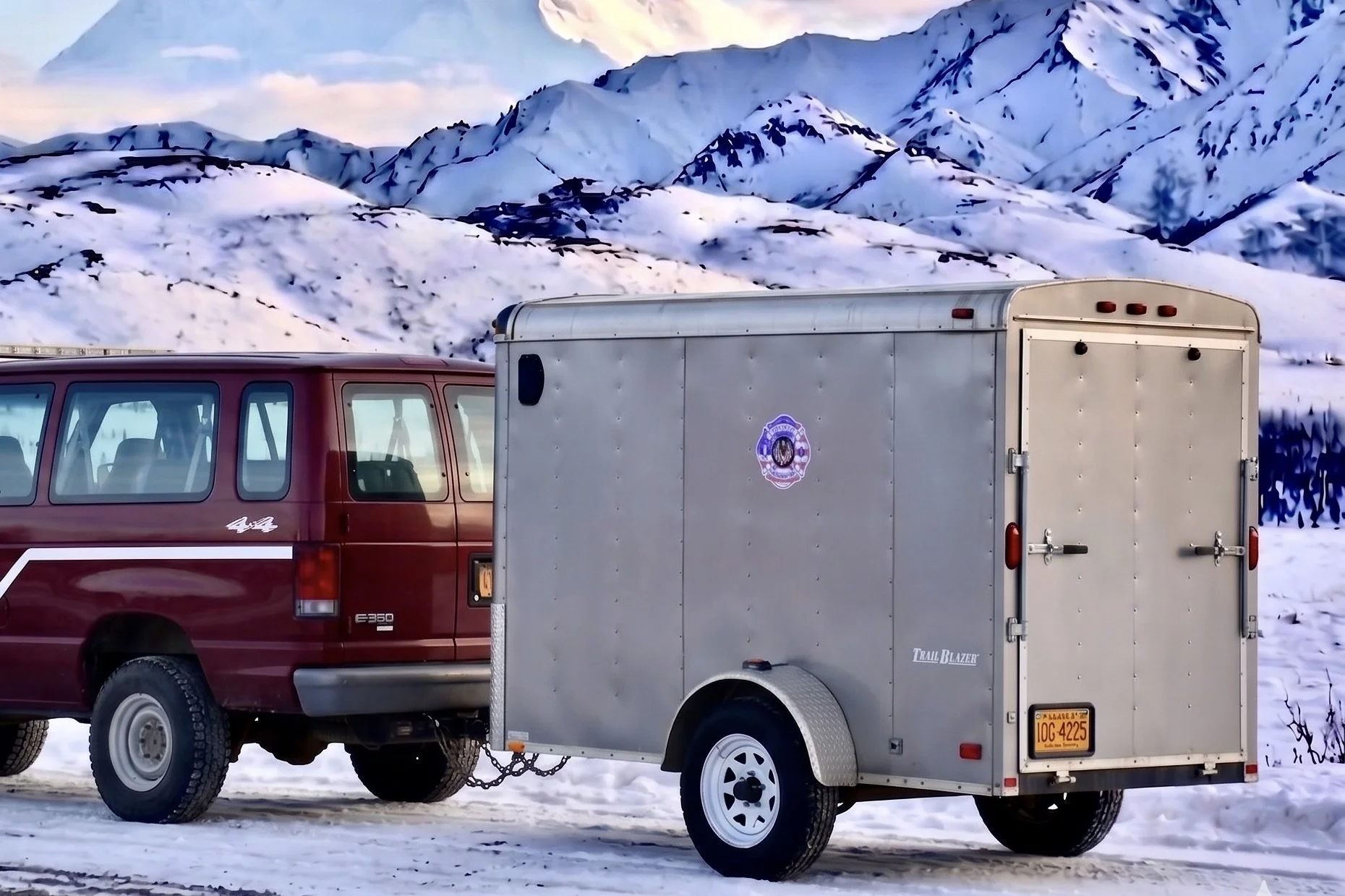 A red vehicle with a utility trailer attached, parked on a snowy landscape with snow-covered mountains in the background.