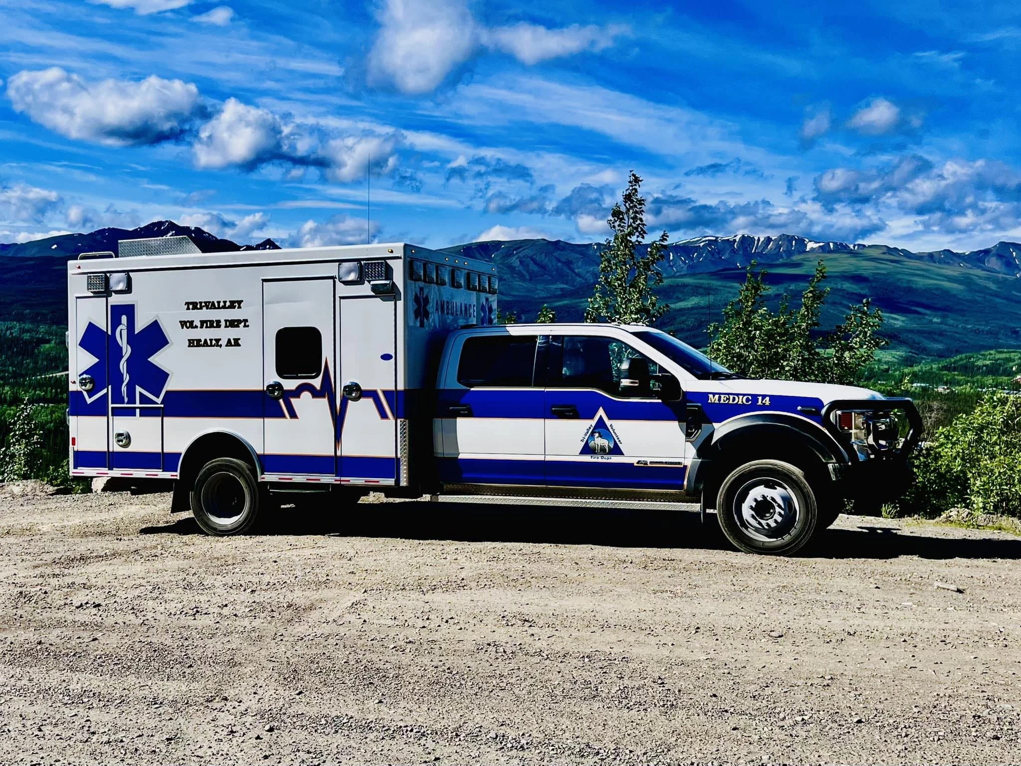 A white and blue ambulance truck parked on a gravel surface with a mountainous landscape and partly cloudy sky in the background.