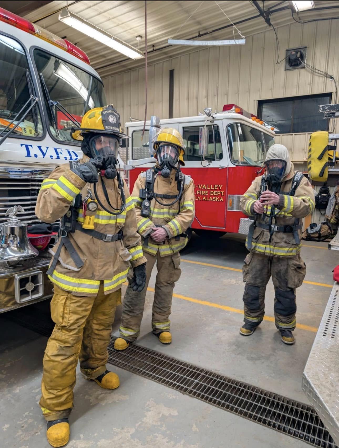 Three firefighters in firefighting gear inside a fire station, with emergency fire trucks behind them.