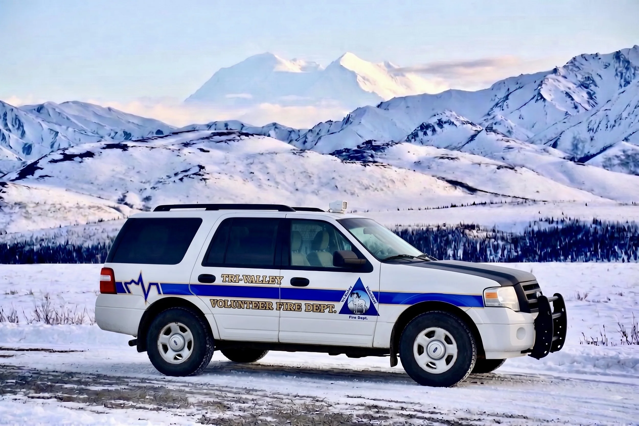 A white Tri-Valley Volunteer Fire Department vehicle parked on snow-covered ground in front of snow-capped mountains with a mountain peak in the distance under a cloudy sky.
