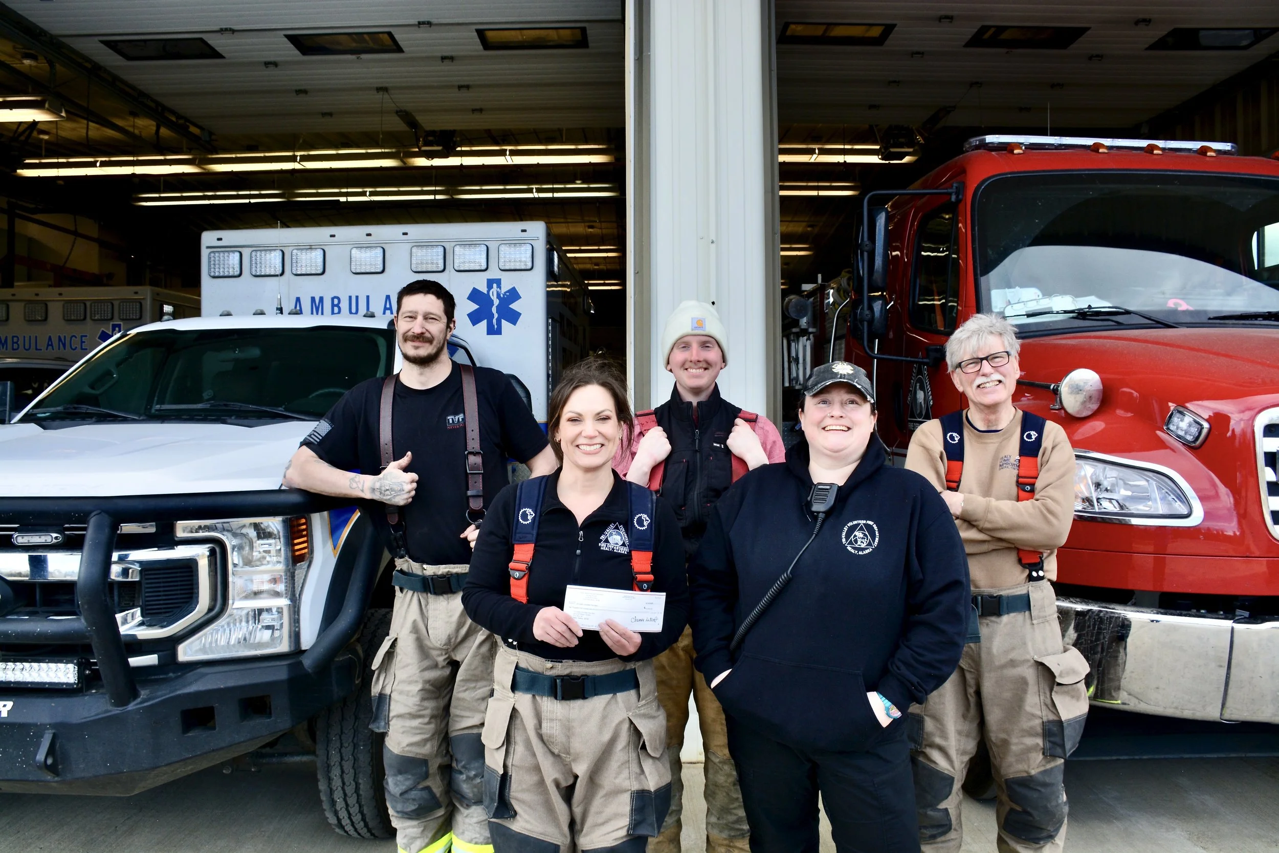 A group of five smiling emergency responders posing in front of an ambulance and fire truck inside a garage.