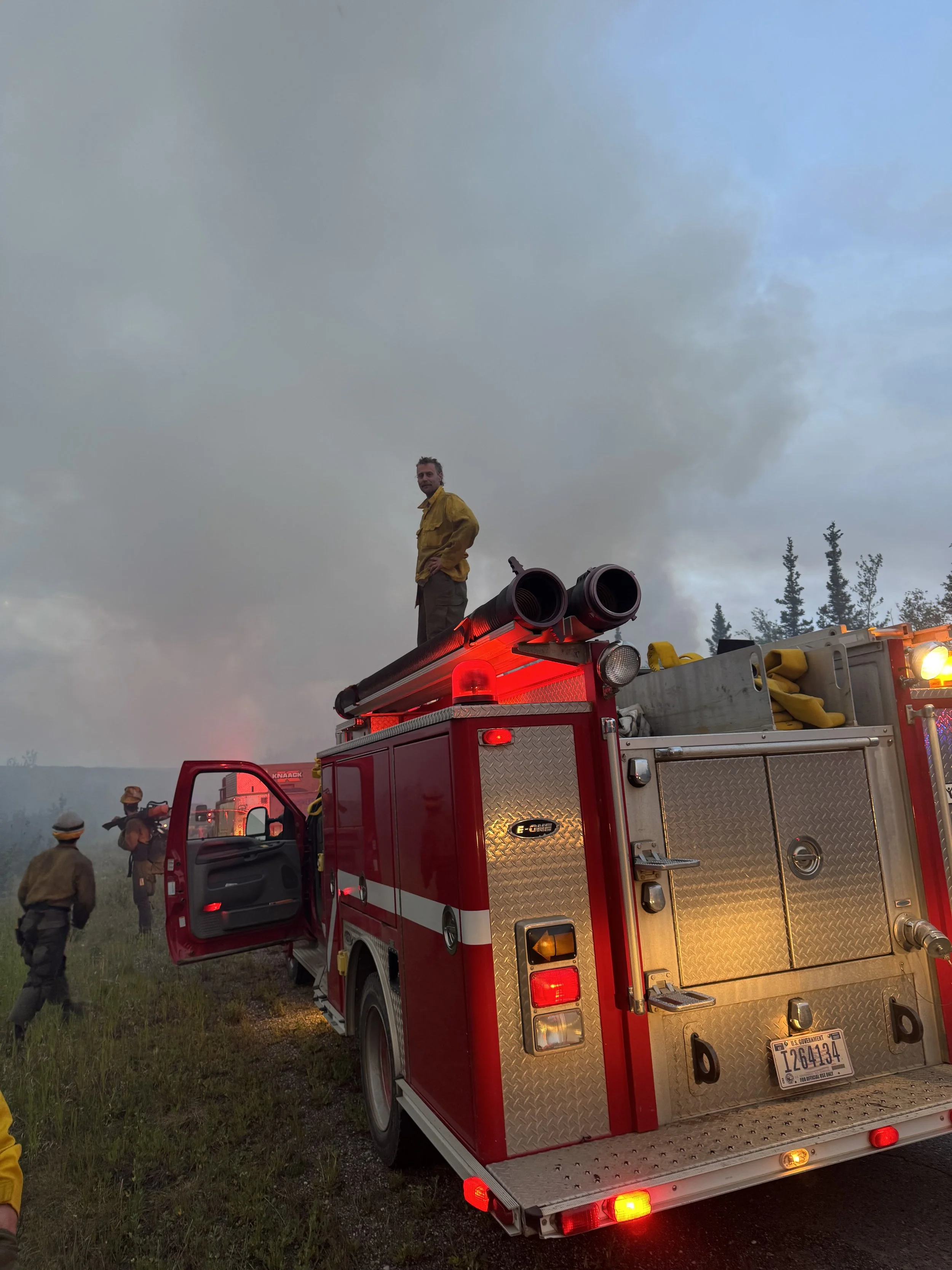 Firefighters on a red fire truck with equipment, standing on a smoky outdoor scene with cloudy sky and trees in the background.