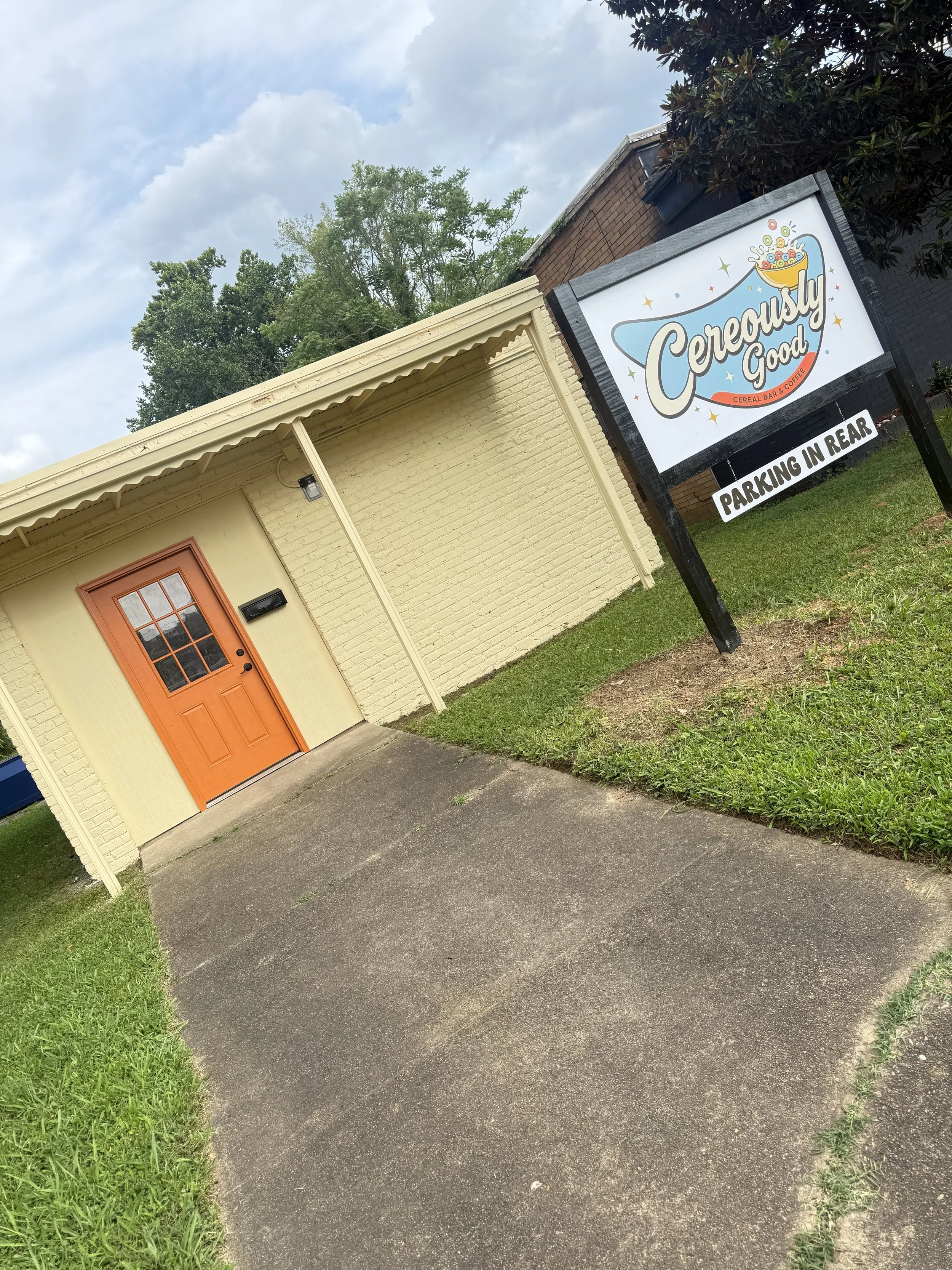 A building with a yellow brick exterior, orange door, and black mailbox, with a large sign that reads 'Cereously Good' and indicates parking in the rear, surrounded by grass and trees under cloudy sky.