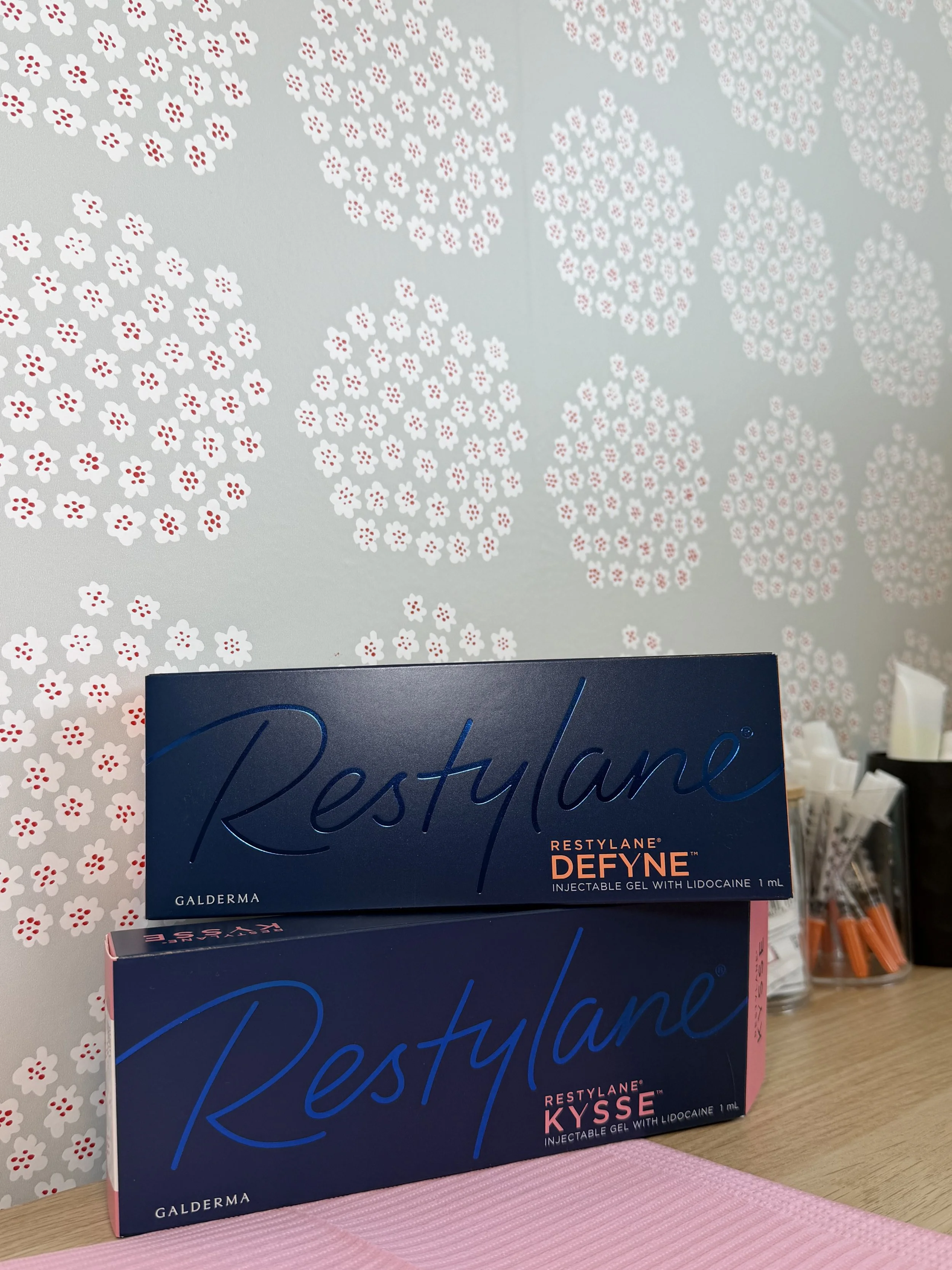 Boxes of Restylane dermal fillers on display, positioned in front of a floral-patterned wall. The boxes are labeled 'Restylane Defyne' and 'Restylane Kysse'.