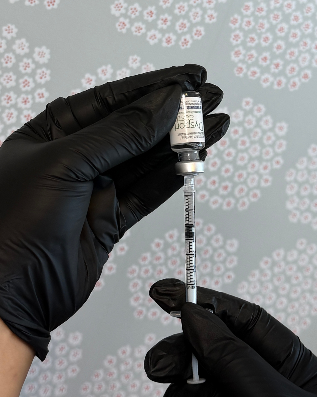 A person wearing black gloves is holding a vaccine vial and a syringe filled with liquid, preparing for administration against a background of patterned wallpaper with red and white flowers.