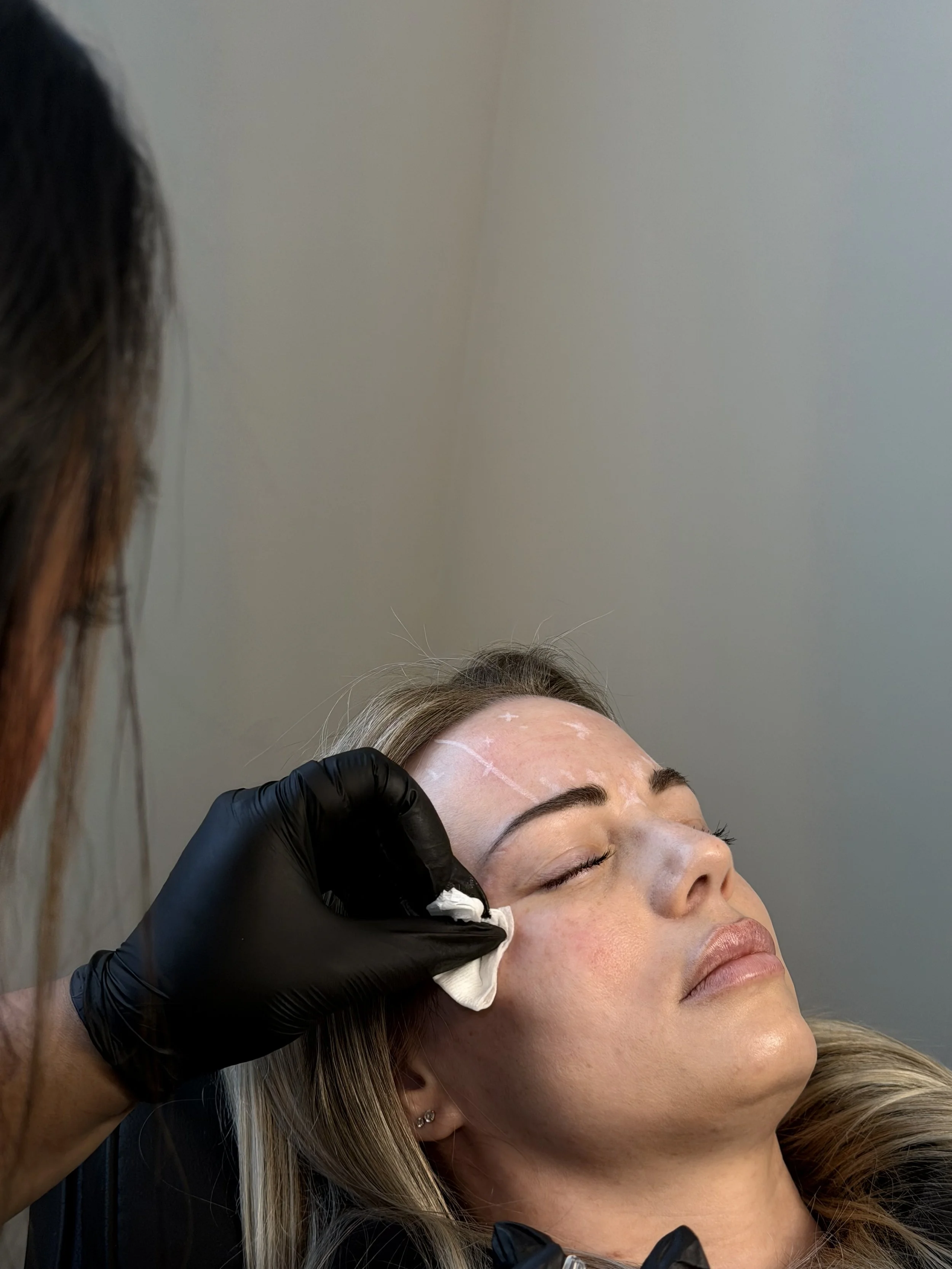 A woman is lying down with her eyes closed, receiving a facial treatment from a professional wearing black gloves. The professional is cleaning or applying a product to the woman's face, which has markings on it. The background is plain and light-colored.