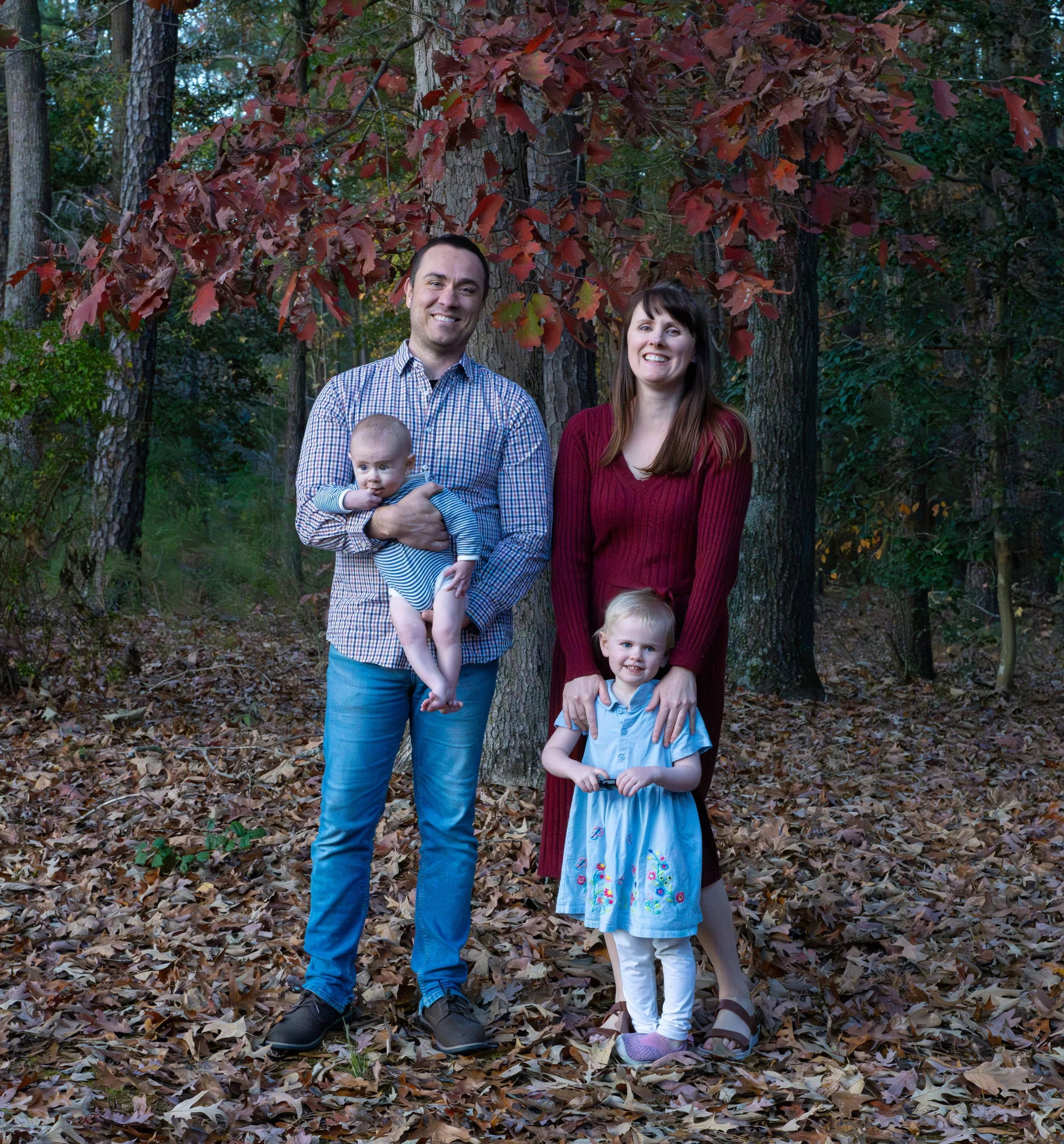 Family of four standing in a wooded area with autumn leaves