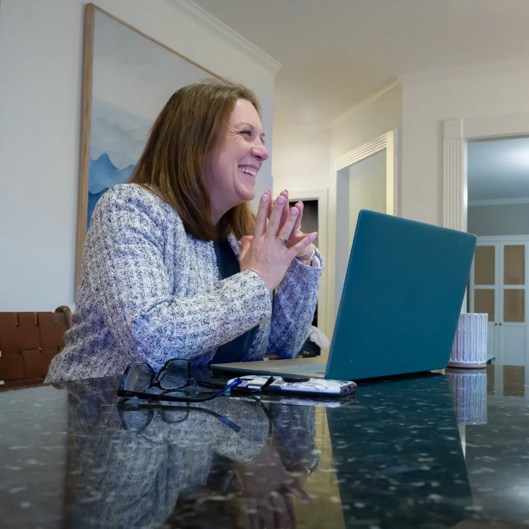 Woman sitting at a table with a laptop, smiling, with glasses and a phone on the table.