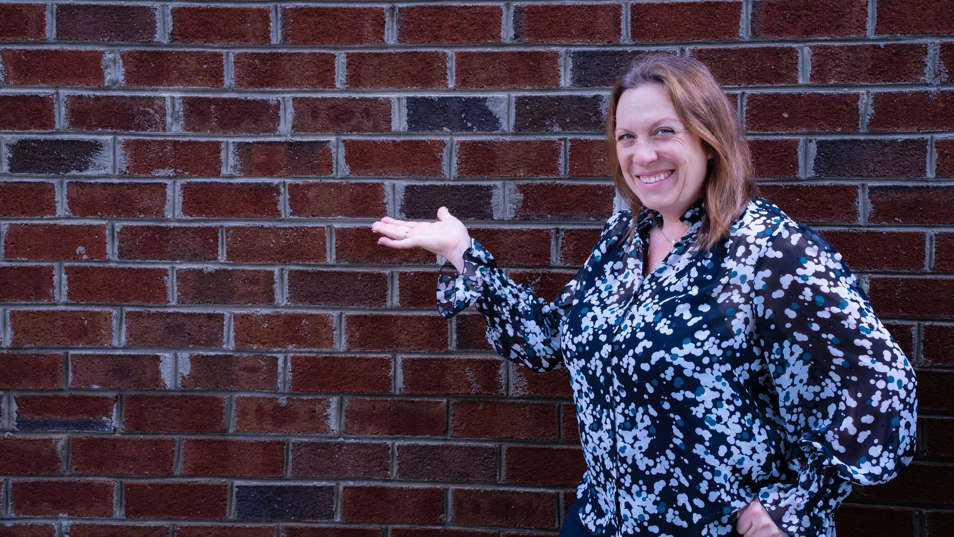 Woman with shoulder-length brown hair, wearing a black blouse with white and blue polka dots, smiling and pointing at a plain brick wall.