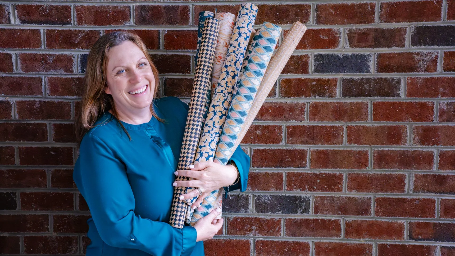 A woman with red hair and blue eyes smiling, wearing a blue shirt, standing in front of a red brick wall, holding several rolls of colorful wrapping paper.