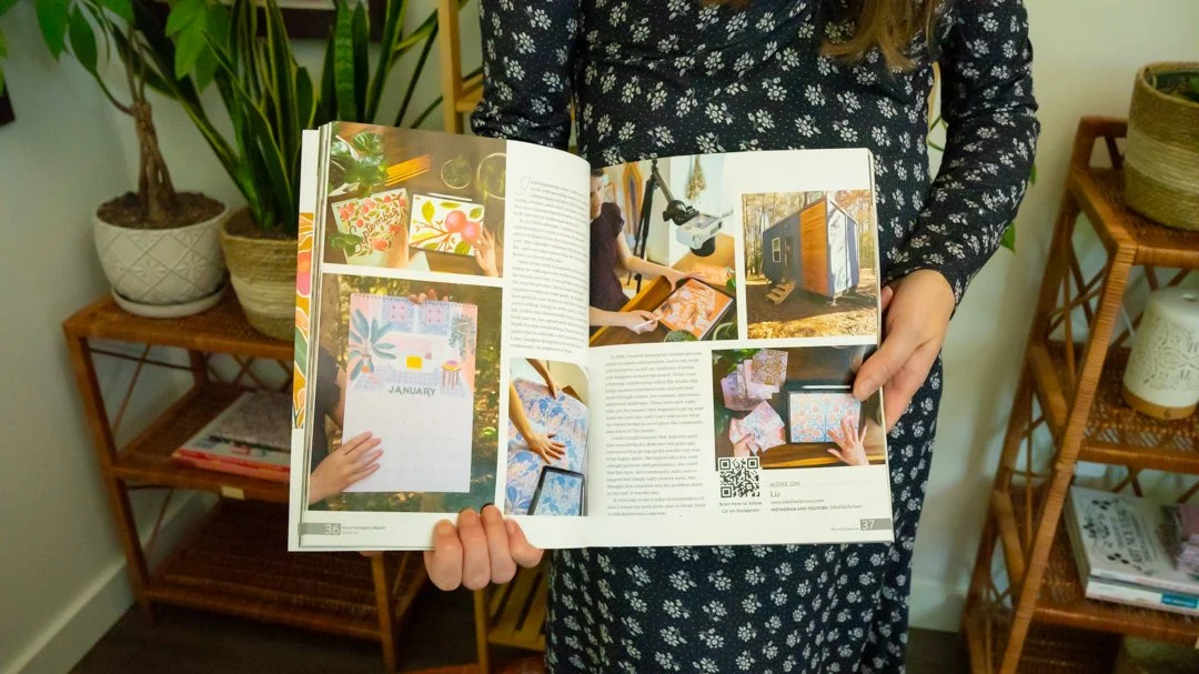 Person holding open magazine featuring craft and art projects displayed on shelves and table in background.