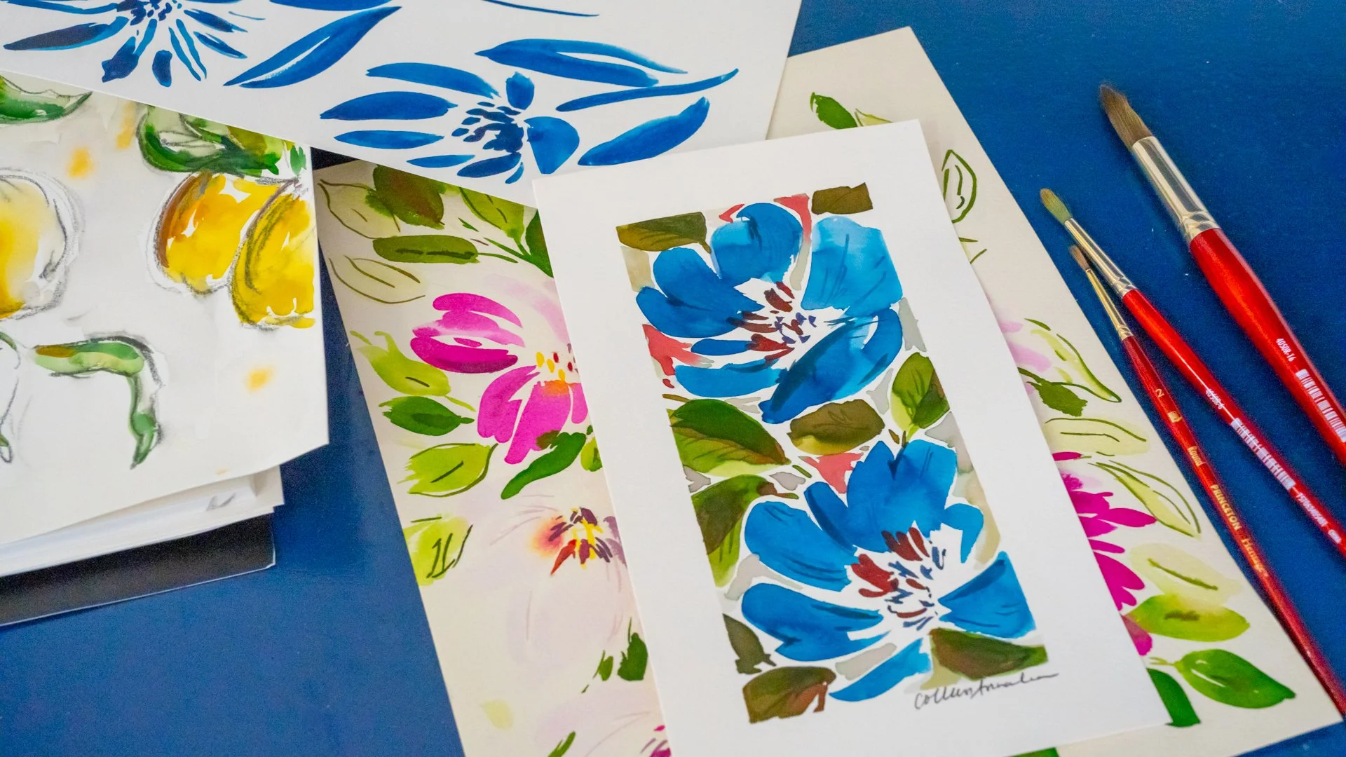 Several watercolor paintings of flowers and leaves in vibrant colors, with brushes and paper on a blue surface.