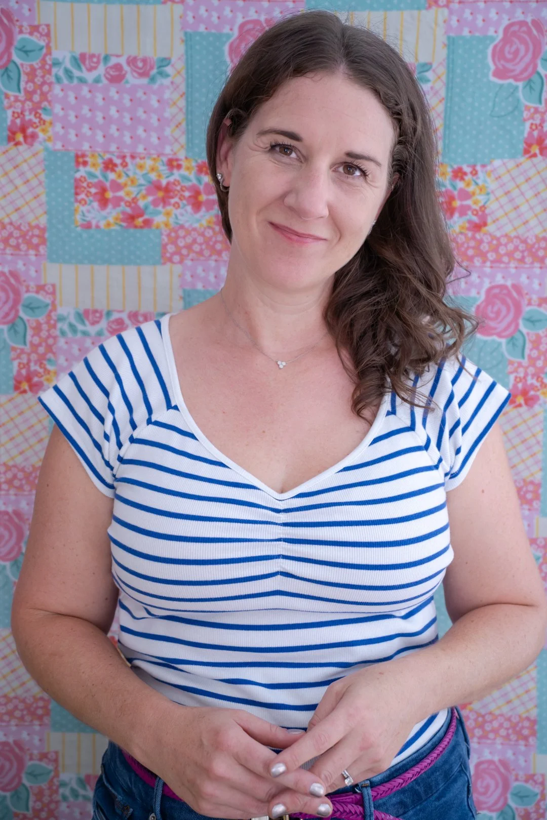 A woman with brown, wavy hair wearing a blue and white striped shirt stands in front of a colorful patchwork quilt with pink, blue, yellow, and floral patterns.