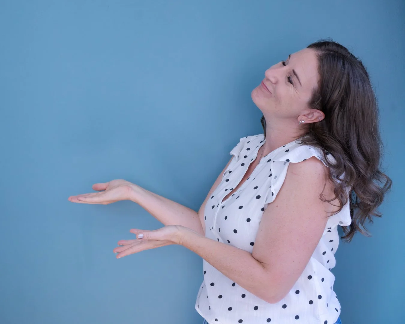 Woman with shoulder-length hair in a white polka dot blouse, smiling and gesturing with both hands against a blue background.