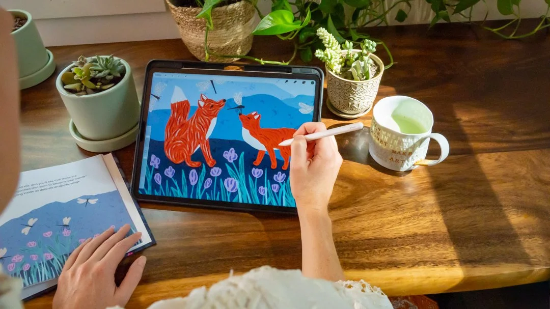 A person drawing digital art of two orange cats in a field of purple flowers on a tablet, surrounded by potted plants and a cup of green tea on a wooden table.