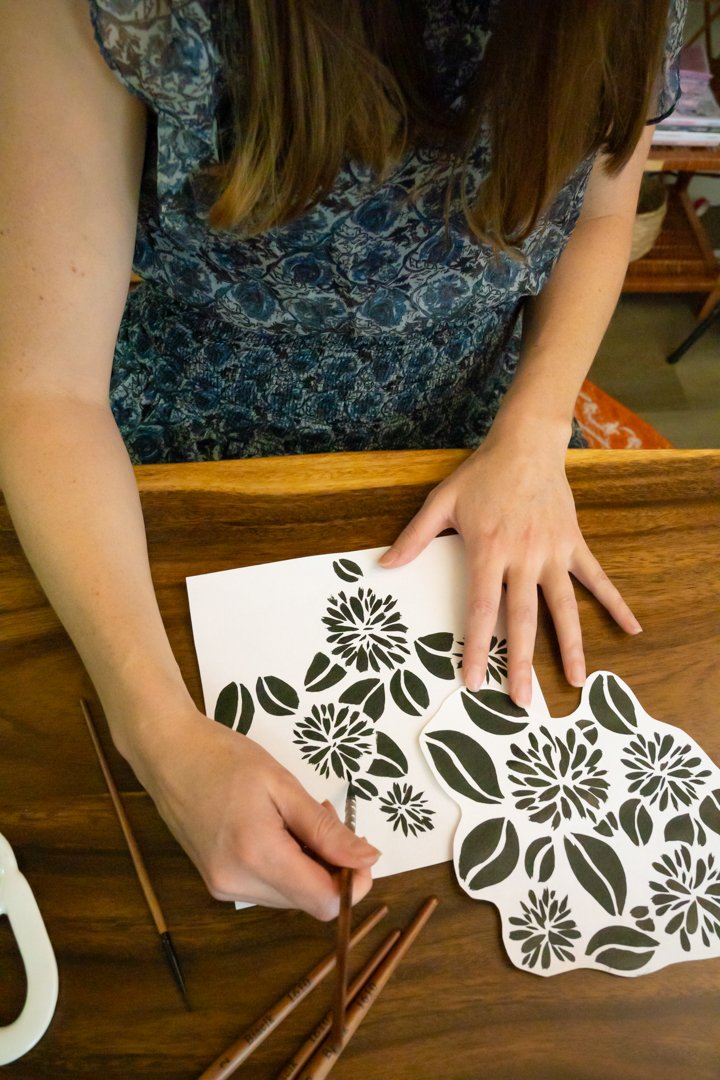Person using paintbrushes to create black floral stencil patterns on white paper.